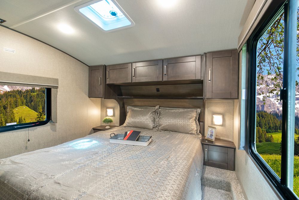Spacious RV rental bedroom featuring a large bed, overhead cabinets, and dual windows showcasing stunning natural views.. Grand Design Reflection 2021