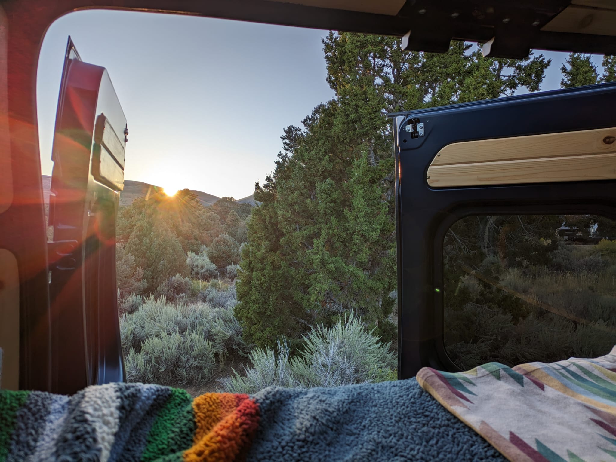 Cozy rv rental view: Sunrise over a natural landscape of trees & sagebrush, seen from inside a camper van's open doors with blankets.. Dodge Ram Promaster 1500 2020