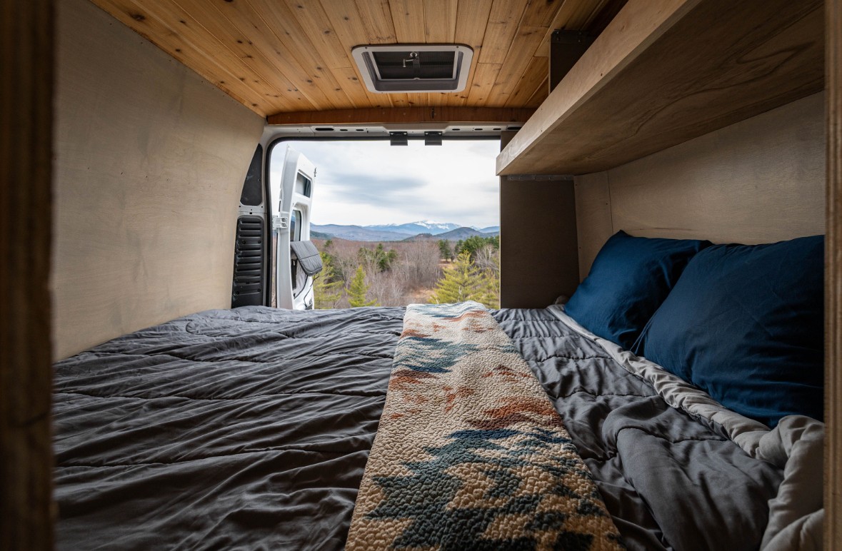 Inside a cozy RV rental: a comfortable bed with a scenic mountain and forest view from the open back doors.. Dodge Sprinter Van 2021