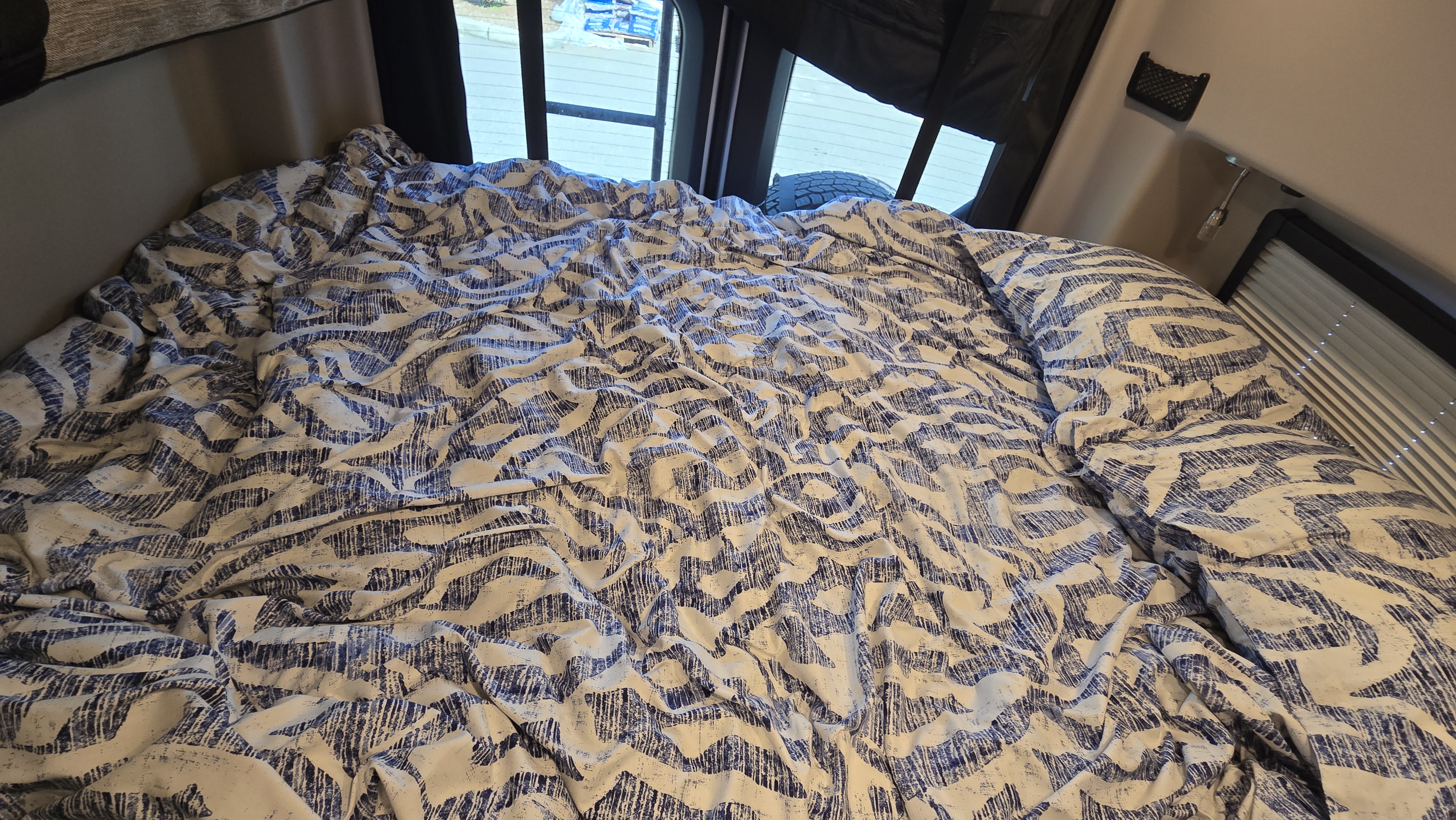 RV rental bedroom: a cozy bed with blue & white patterned bedding, visible rear windows, and side blinds.. Entegra Coach Launch 2023