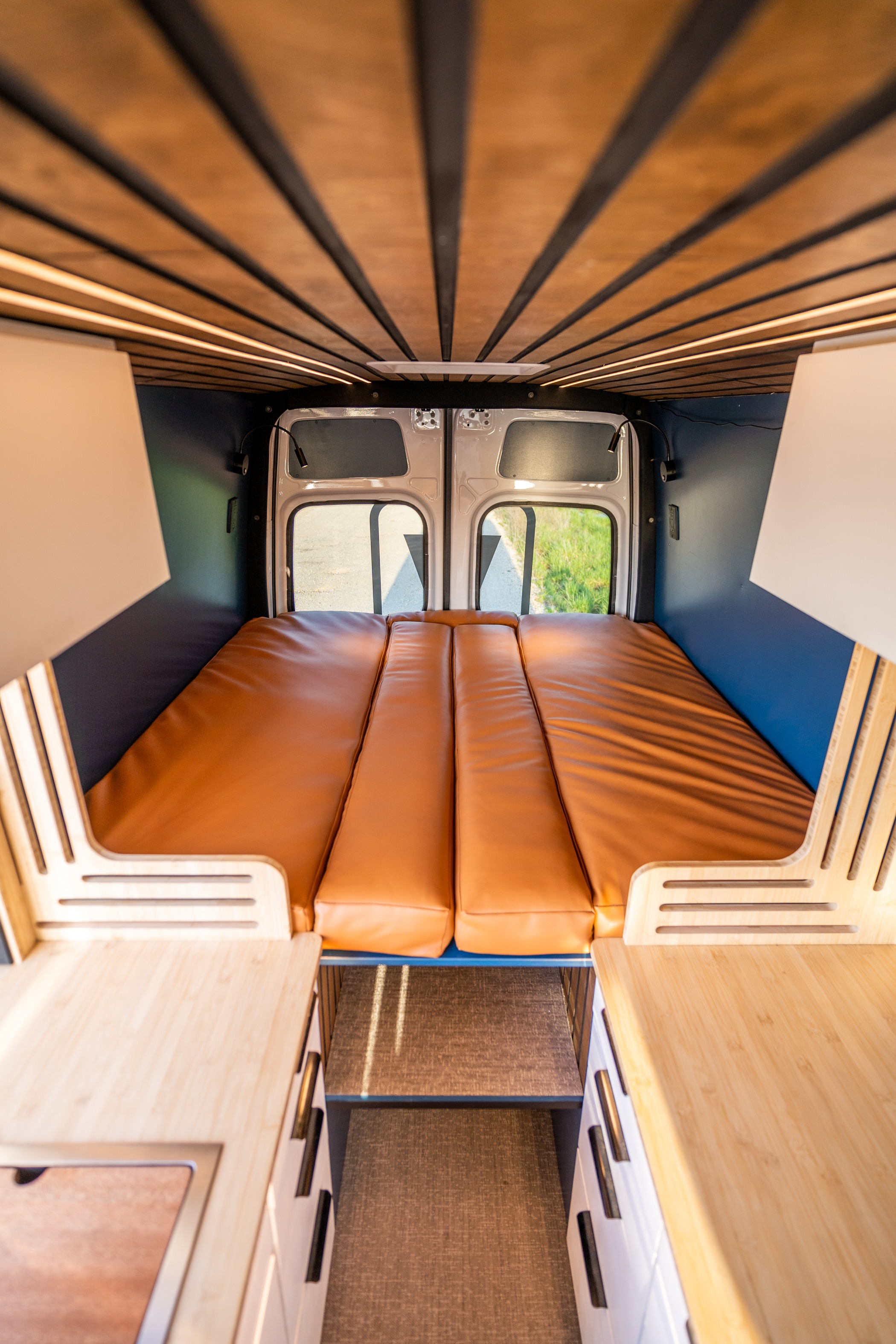 An inviting rv rental interior with a slatted wooden ceiling, warm lighting, a plush brown bed, and light wood storage cabinets.. Mercedes-Benz Sprinter 2024
