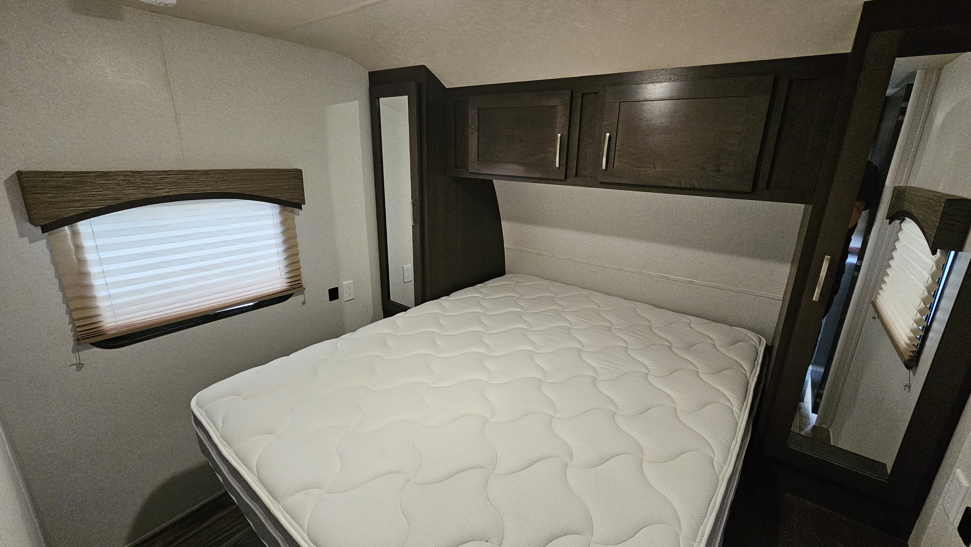 RV rental bedroom with a white quilted mattress, dark wood overhead cabinets, and a window with blinds.. Forest River Evo 2017