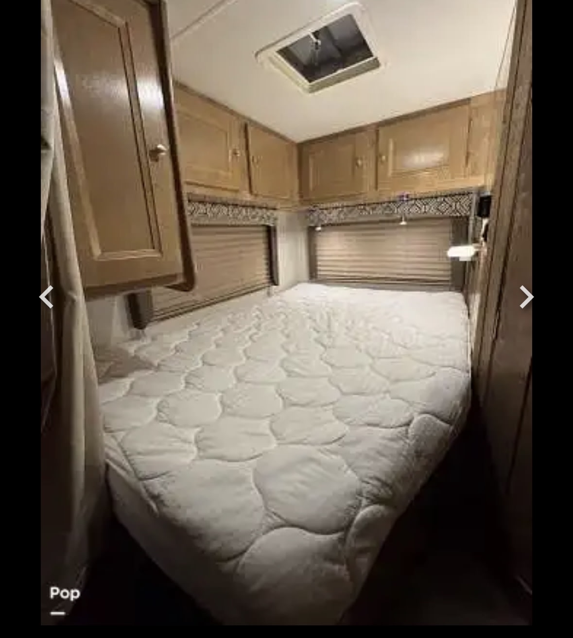 View of an RV rental bedroom with a large bed, overhead wood cabinets, window blinds, and a roof skylight.. Coachman Leprechaun 2020