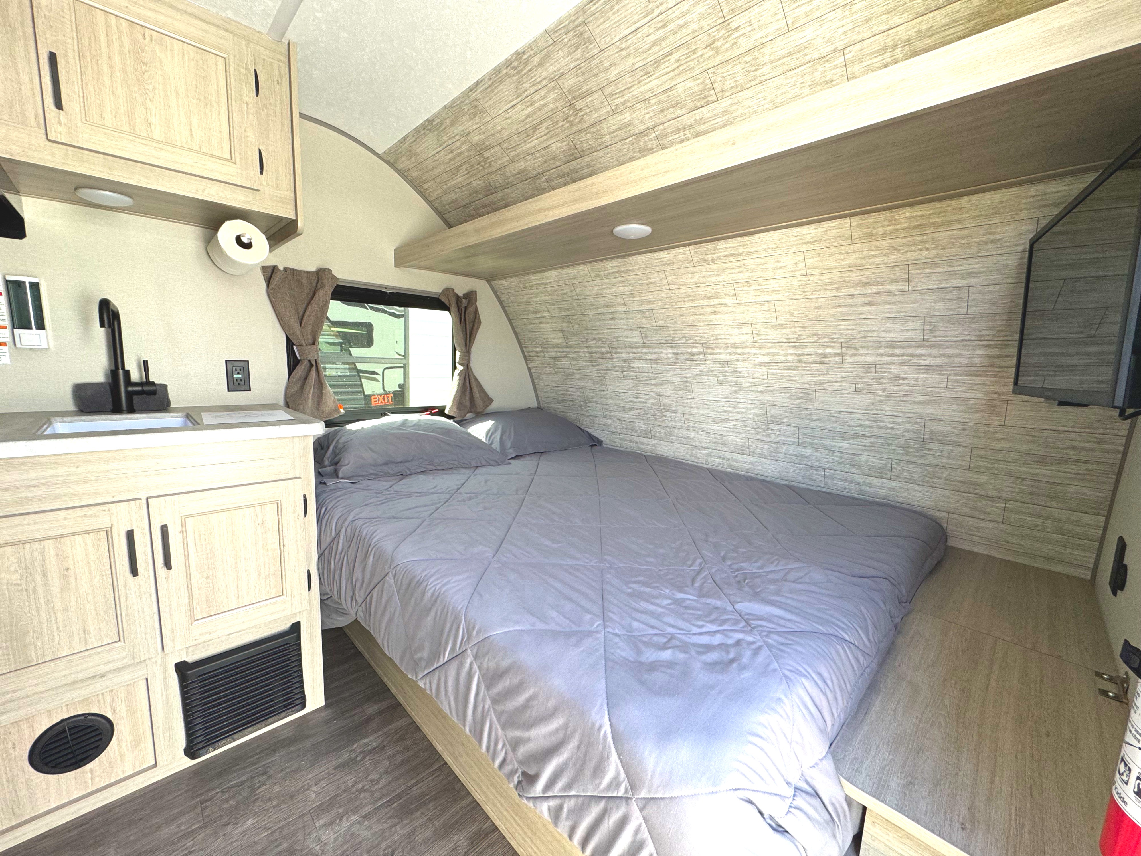 Cozy interior of an RV rental featuring a comfortable bed, kitchenette with sink, and overhead storage. Ideal for travel.. Forest River Aurora 13B 2025