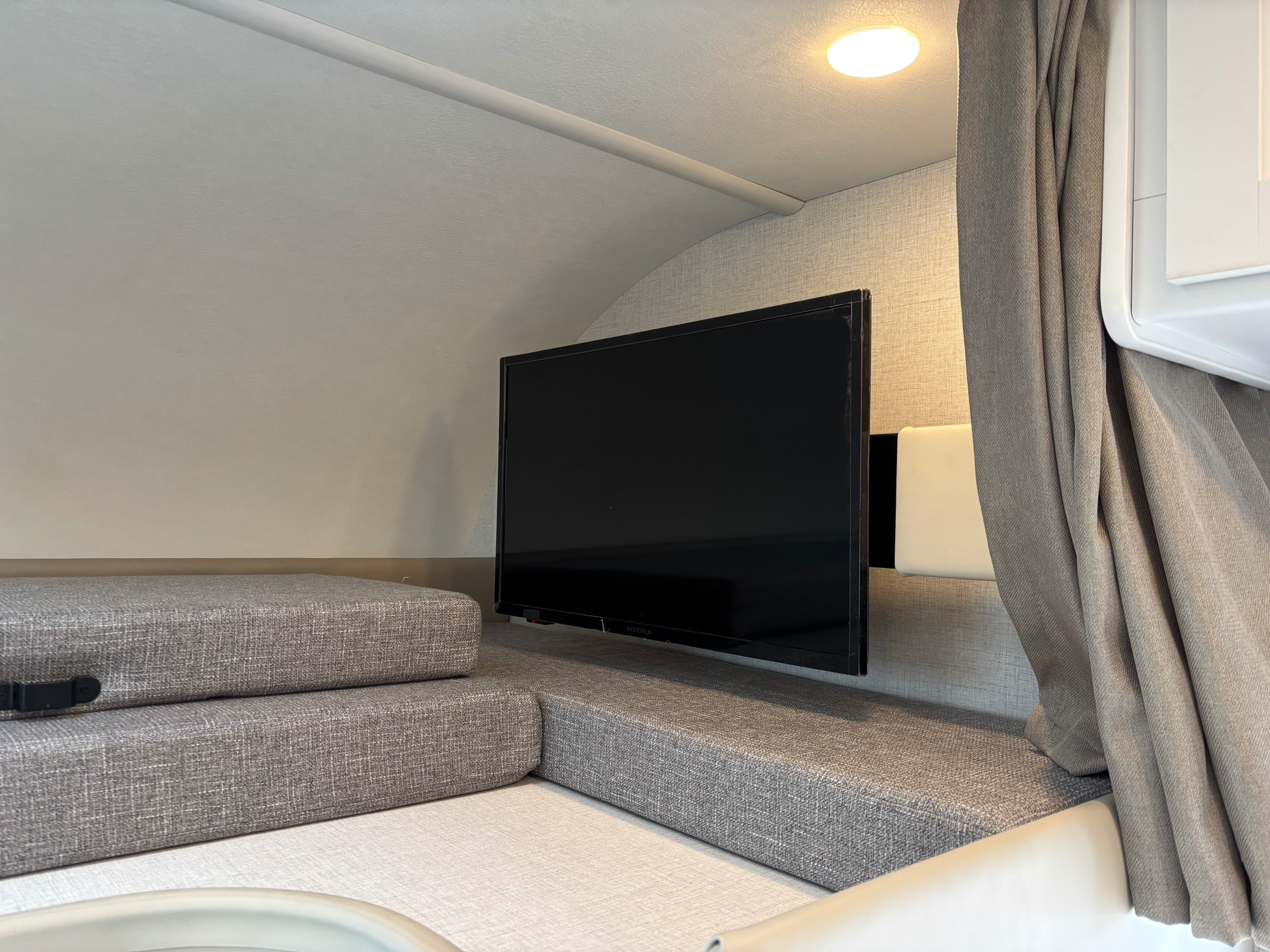 RV rental interior: wall-mounted TV, gray upholstered seating, overhead light, and privacy curtain. A compact entertainment setup.. Thor Motor Coach Four Winds 2026