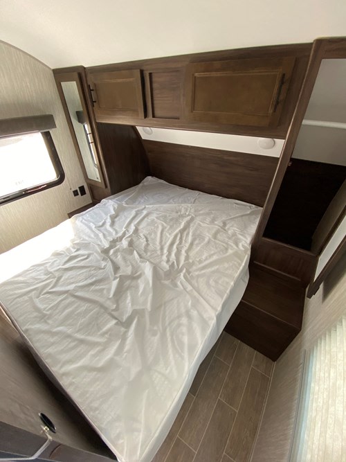 This RV rental bedroom features a bed, dark wood overhead cabinets, a side window, and a built-in storage nook.. Cherokee Grey Wolf 2021