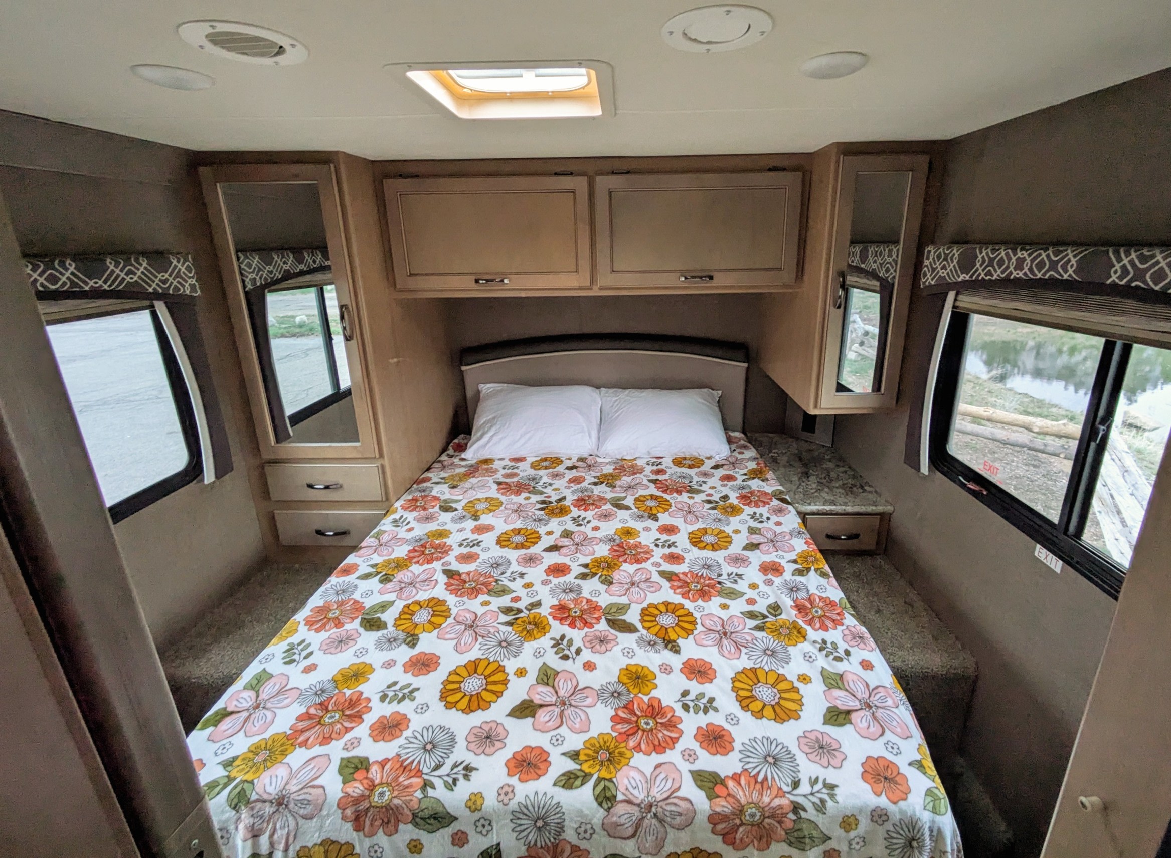 Cozy RV rental bedroom featuring a floral bed, overhead storage, and windows with scenic views.. Thor Chateau 2018