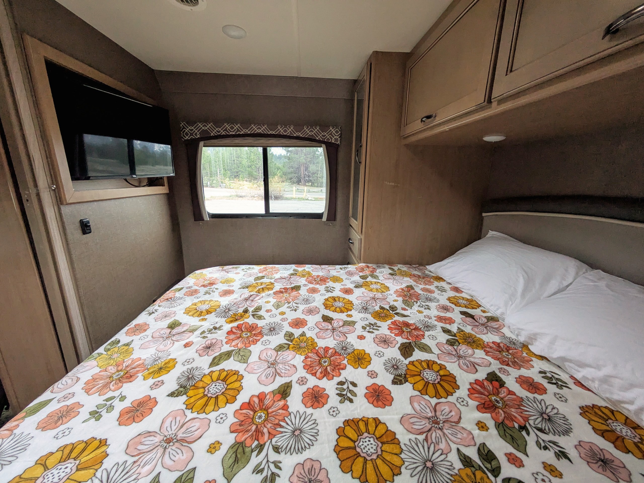 Inside a comfy RV rental bedroom: floral bed, TV, and window with forest scenery.. Thor Chateau 2018