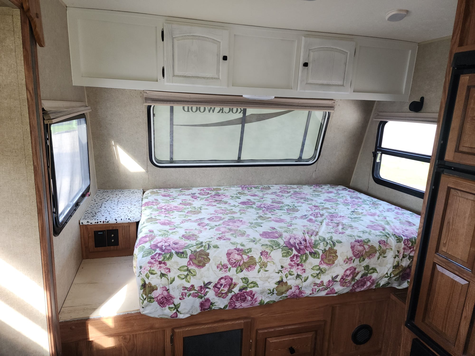 Comfortable RV rental bedroom with a floral bedspread, white overhead cabinets, and windows on three sides.. Forest River Rockwood Mini Lite 2014