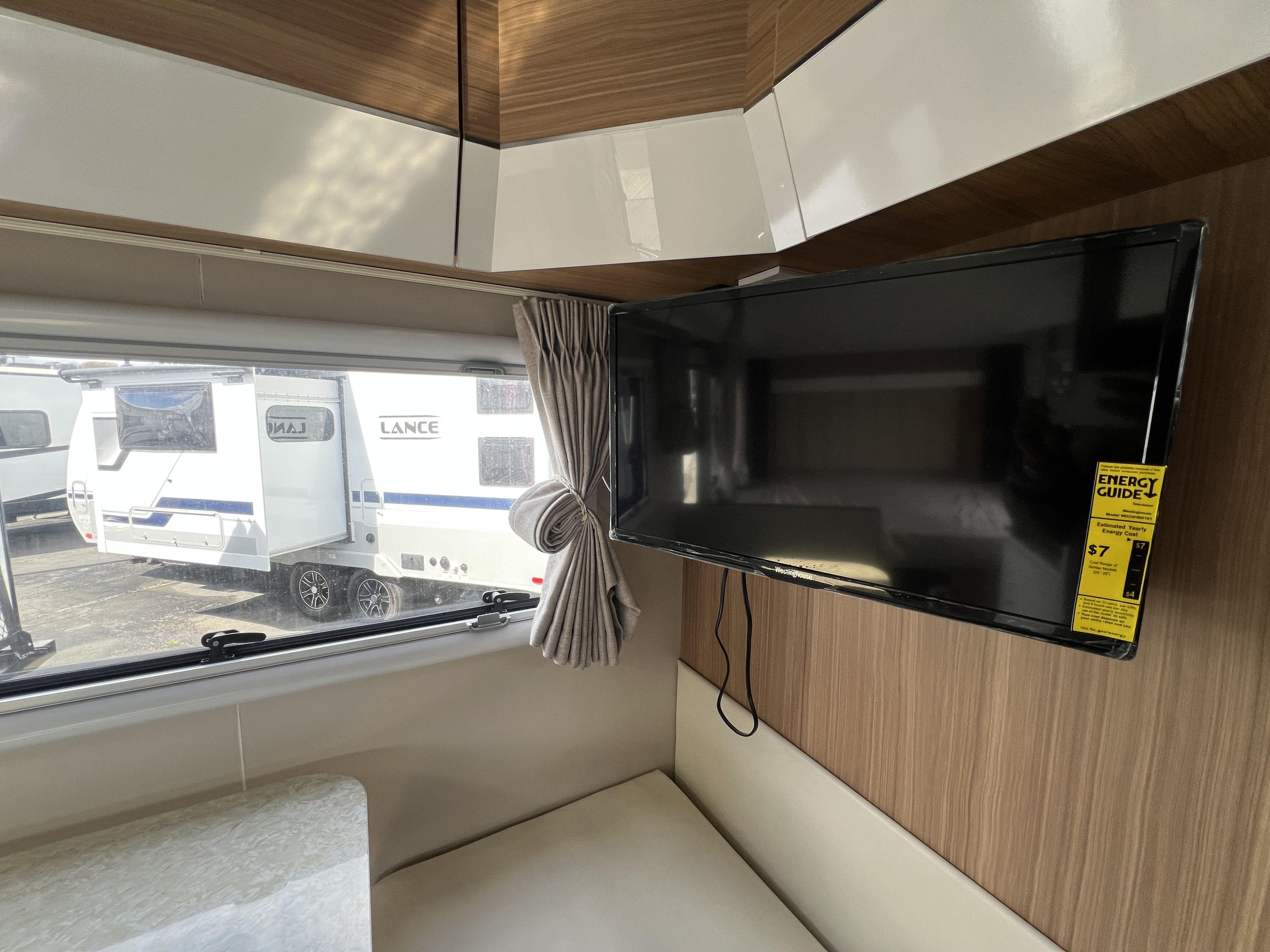 View inside a modern RV rental, showing a TV, curtain, and seating, with a Lance camper visible through the window.. Black Series HQ 19 2025