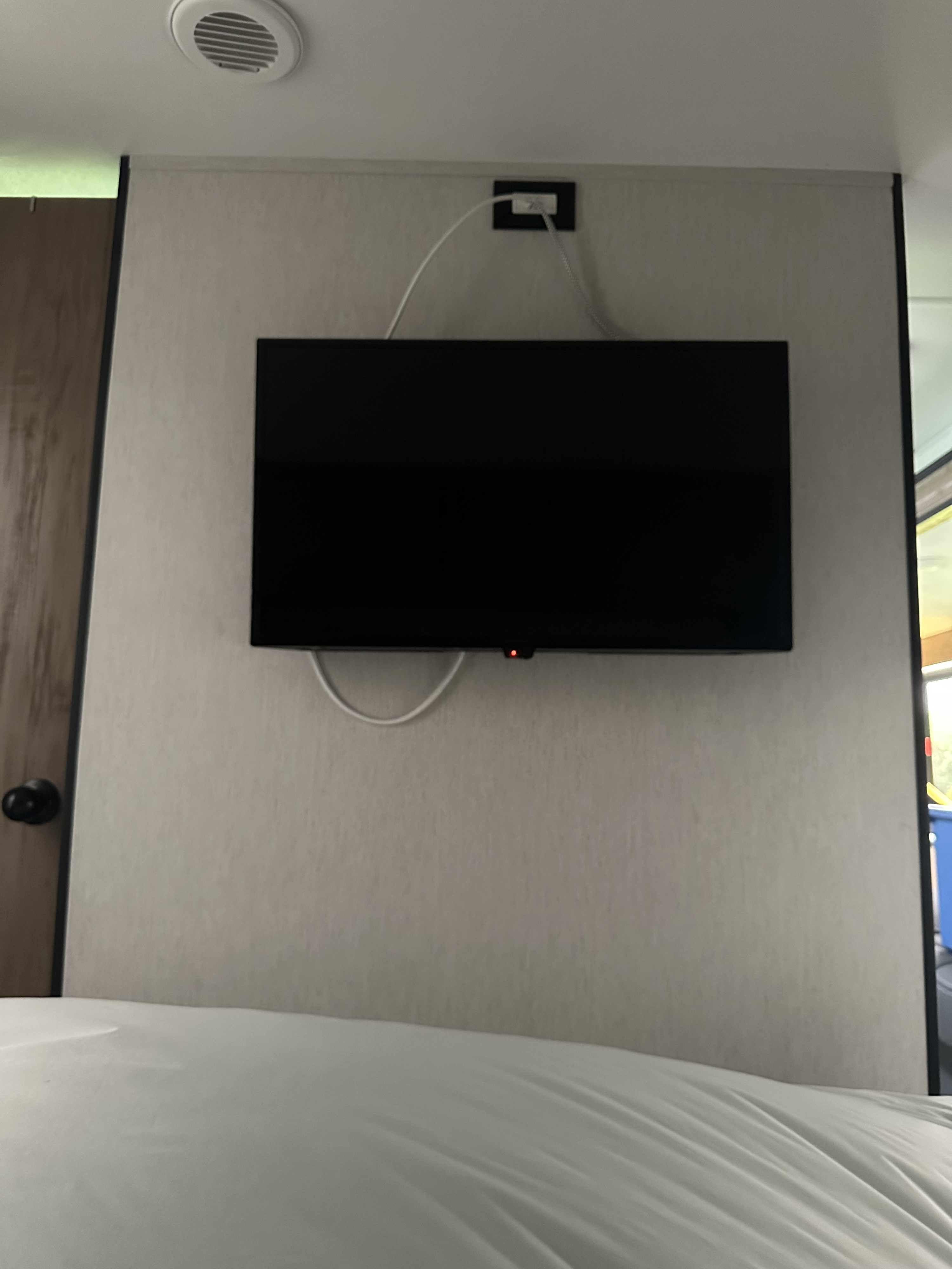 RV rental interior: Wall-mounted TV with red light, power cable, and outlet, above a white bed.. Forest River Cherokee 2023