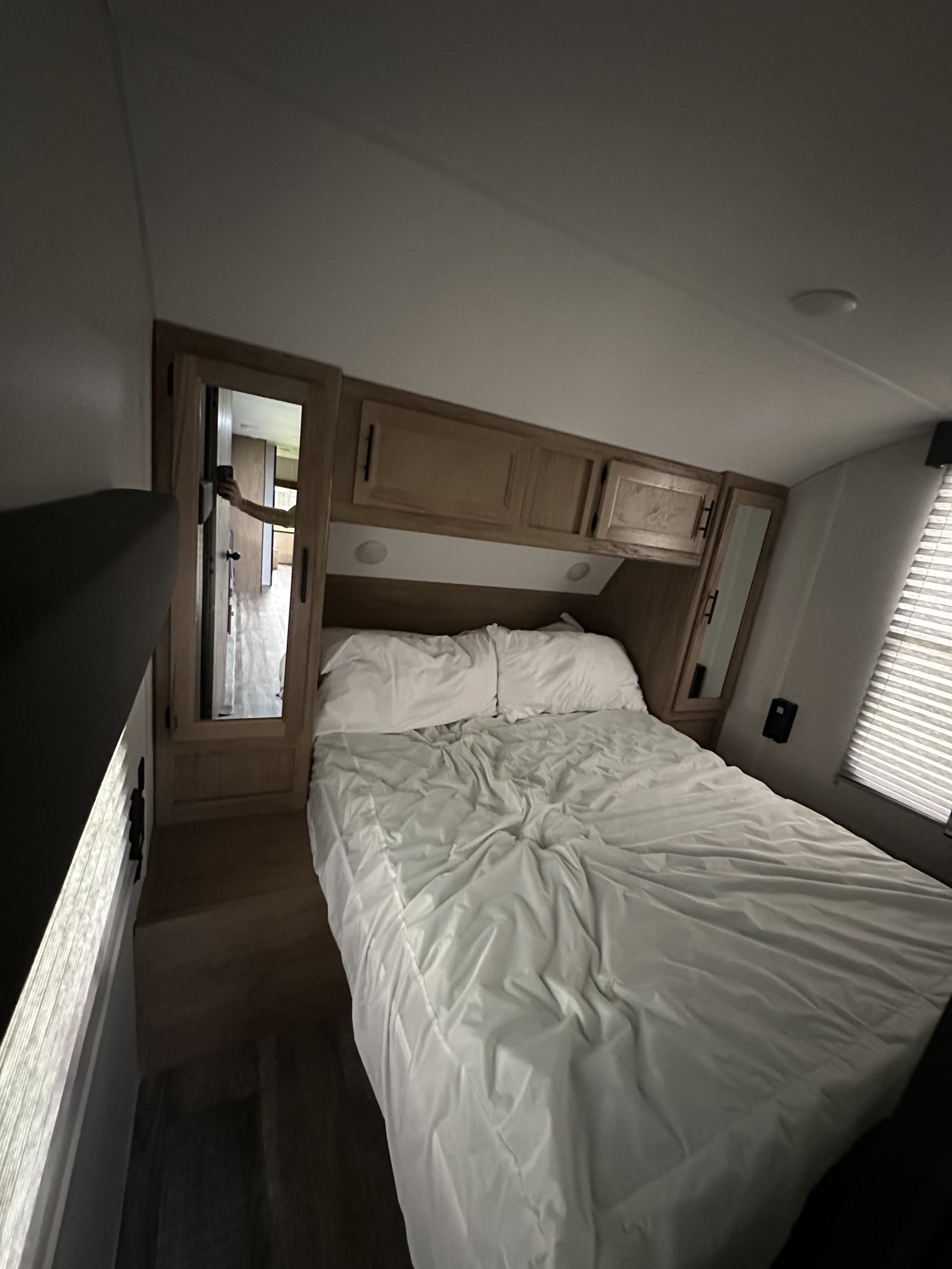 Inside an RV rental, a compact bedroom with a white-sheeted bed, wooden overhead cabinets, and mirrored doors.. Forest River Cherokee 2023