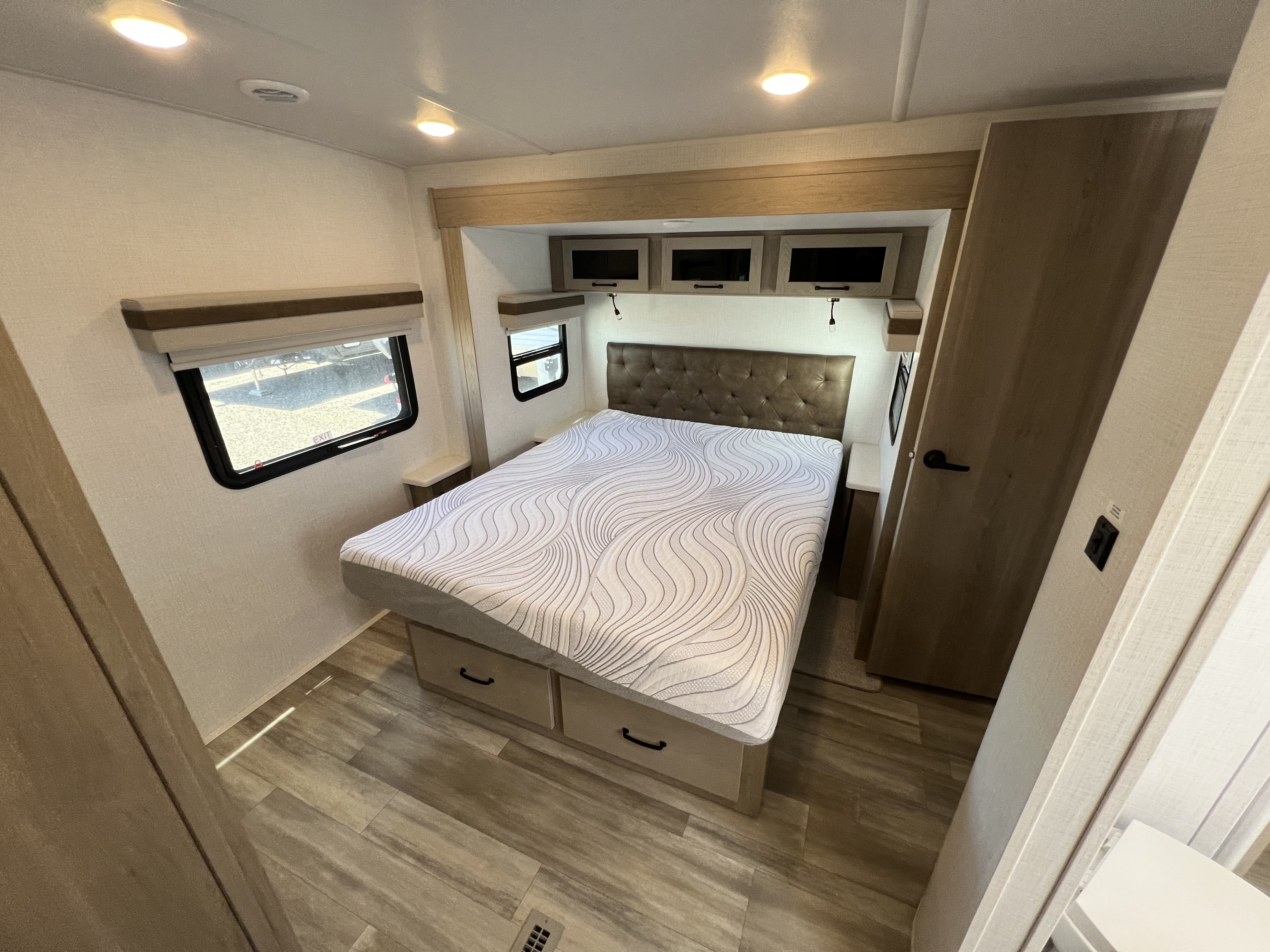 Cozy RV rental bedroom featuring a patterned mattress, tufted headboard, and wood-look flooring, with windows and overhead storage.. Forest River Rockwood Ultra Lite 2024