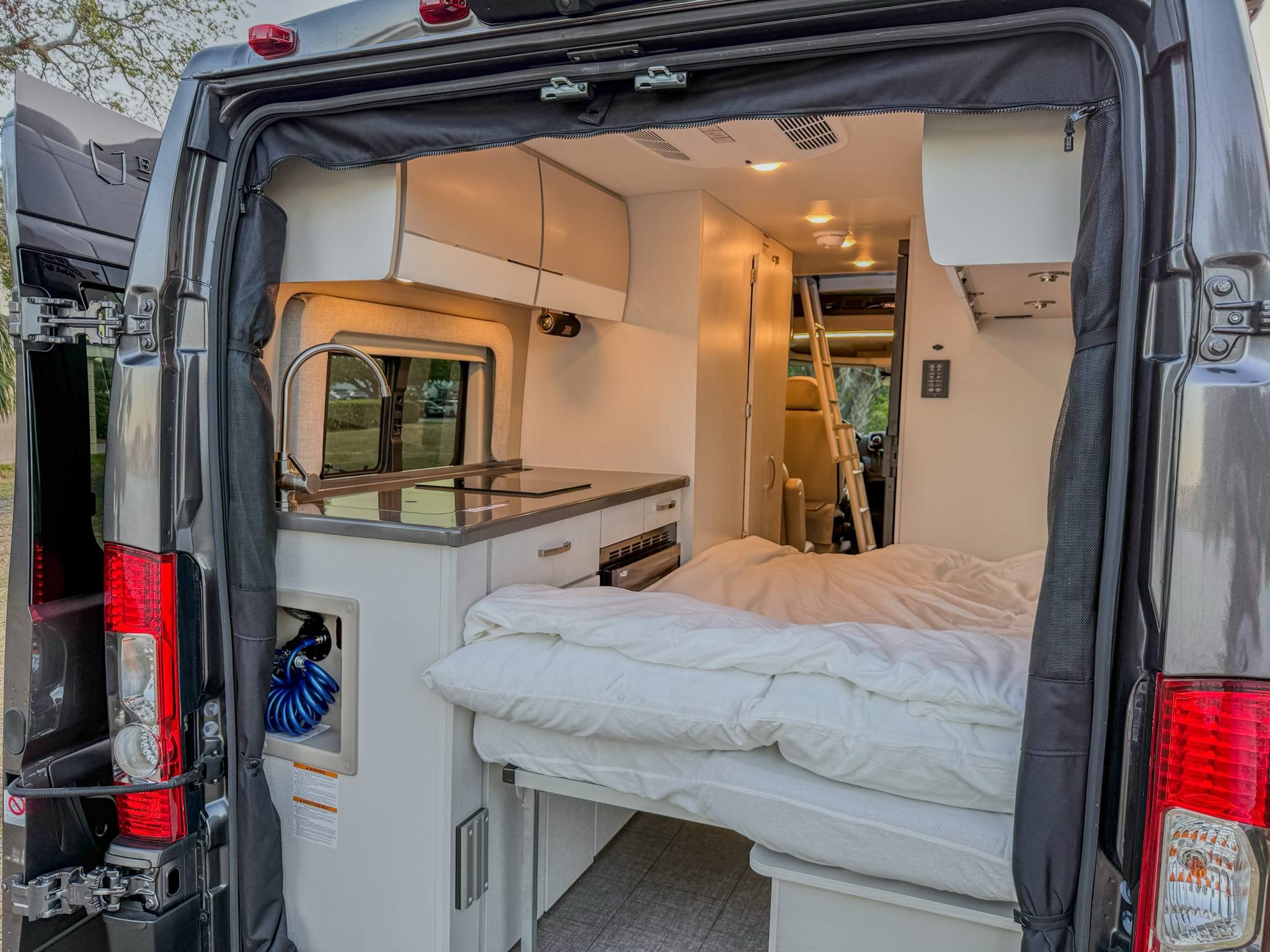 This RV rental interior reveals a compact camper van with a mobile kitchen, sink, and comfortable bed, viewed from the open rear doors.. ENTEGRA ETHOS 2025