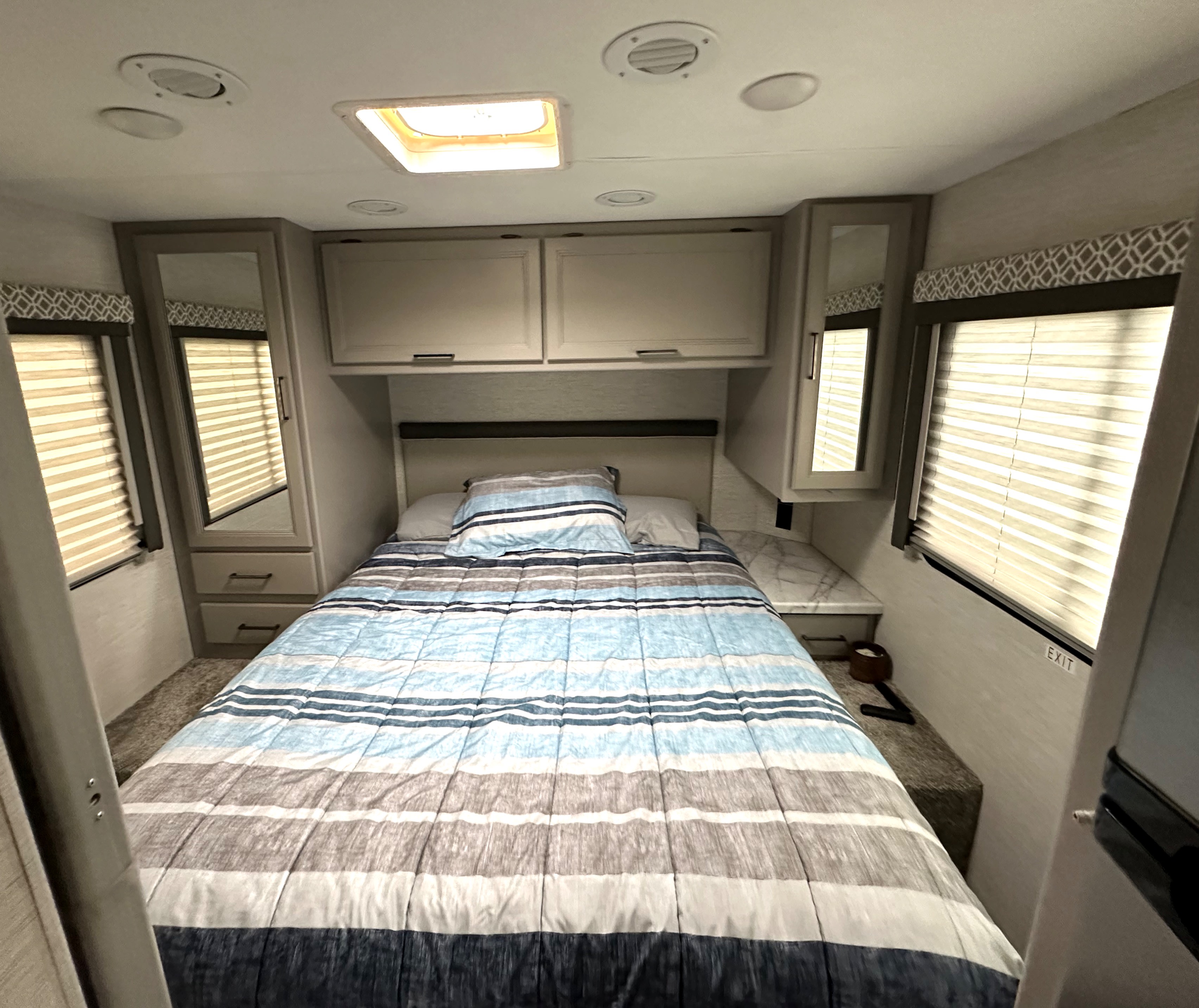 Cozy RV rental bedroom with striped bedding, overhead cabinets, and windows with blinds, ideal for travel.. Thor Coleman 2024