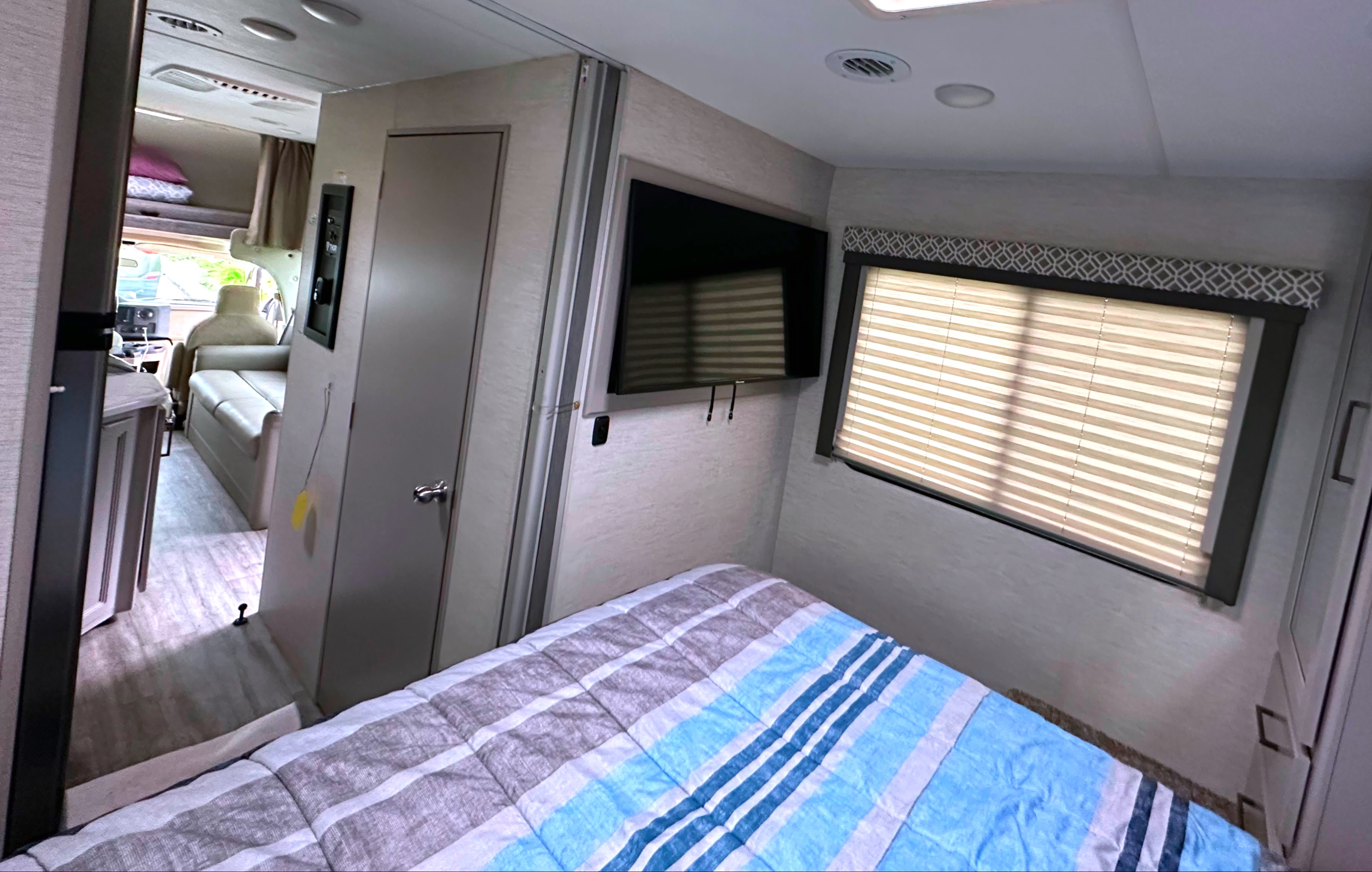An inviting RV rental interior shows a striped bed, wall-mounted TV, window, and hallway leading to the front cab.. Thor Coleman 2024