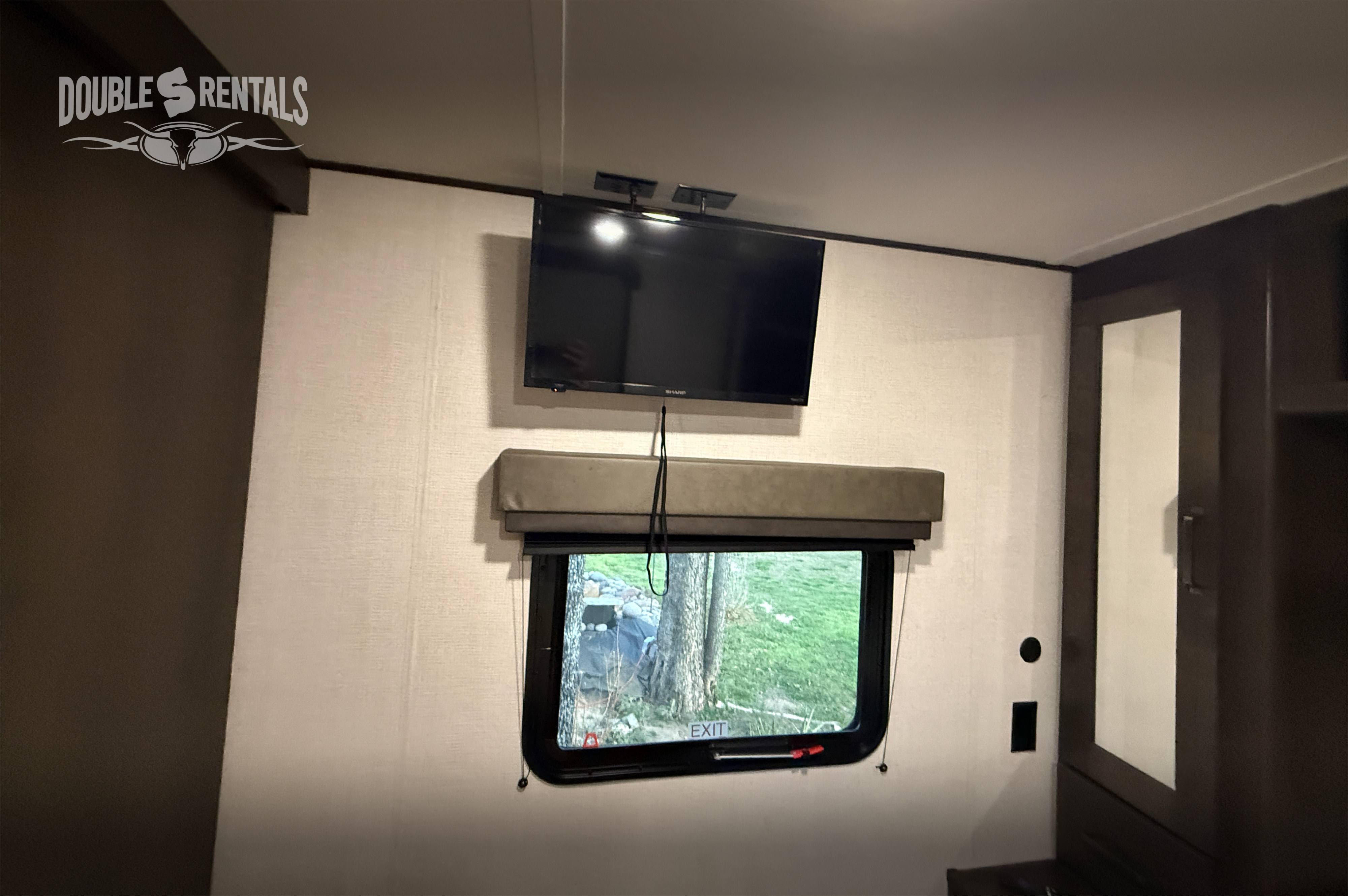 Inside this RV rental: a wall-mounted TV and window showing outdoor trees and grass. Features include dark cabinetry.. Grand Design Transcend 2023