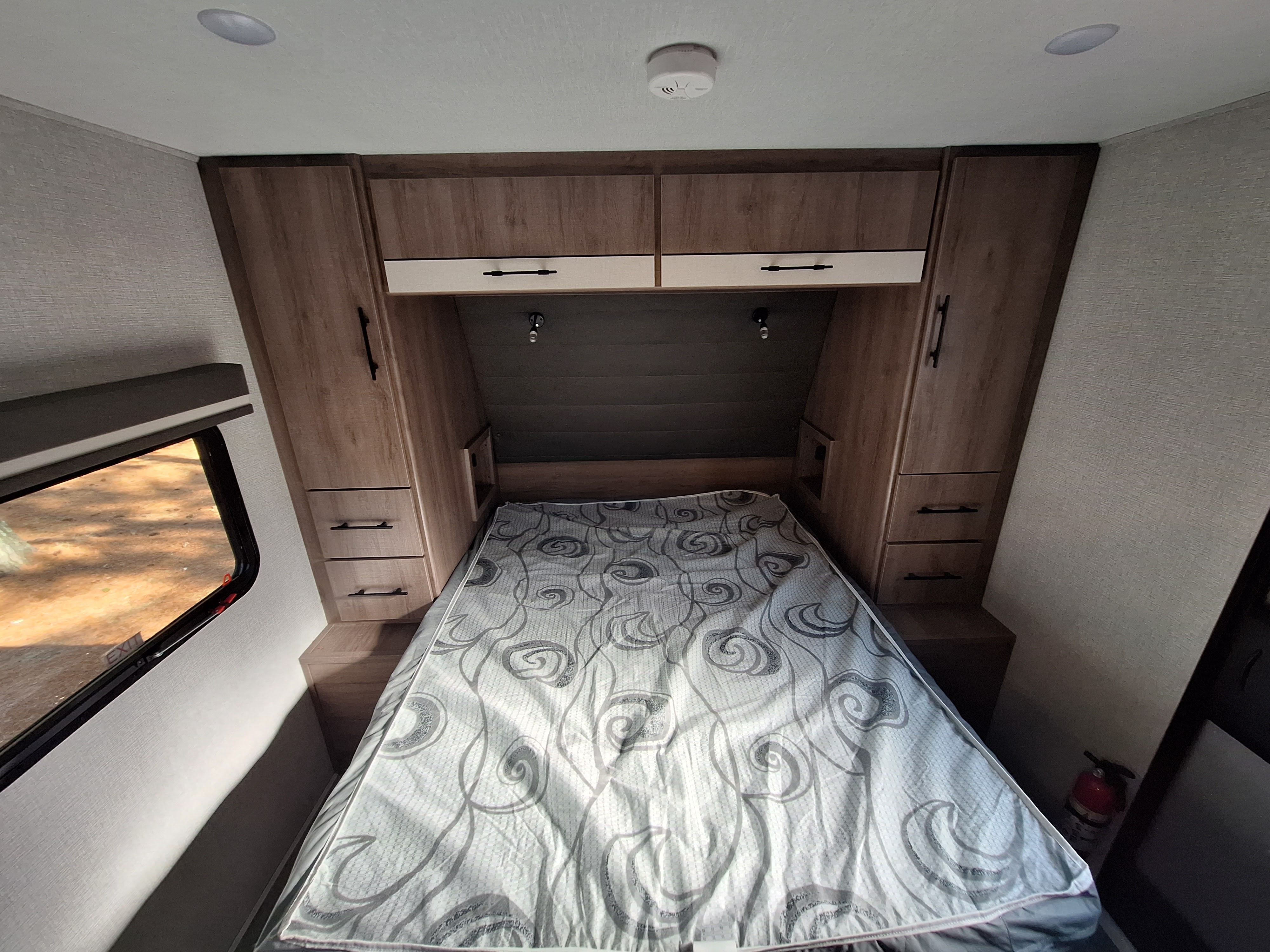 RV rental bedroom with patterned bed, overhead cabinets, and essential safety features like a smoke detector and fire extinguisher.. Grand Design Imagine XLS 2023