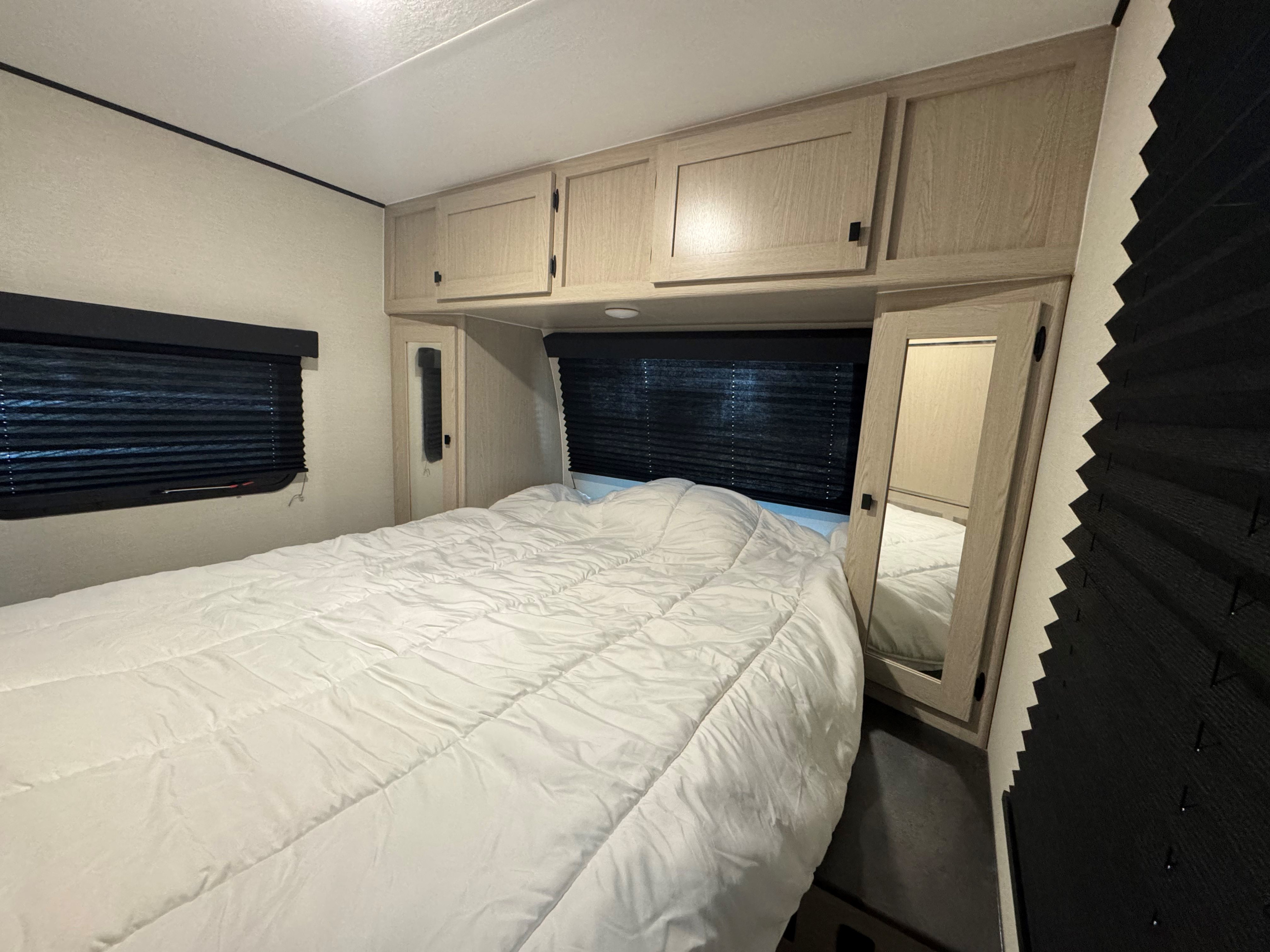 RV rental bedroom with a comfy white bed, overhead wooden cabinets, and dark window blinds for privacy.. Coachmen Apex 2024