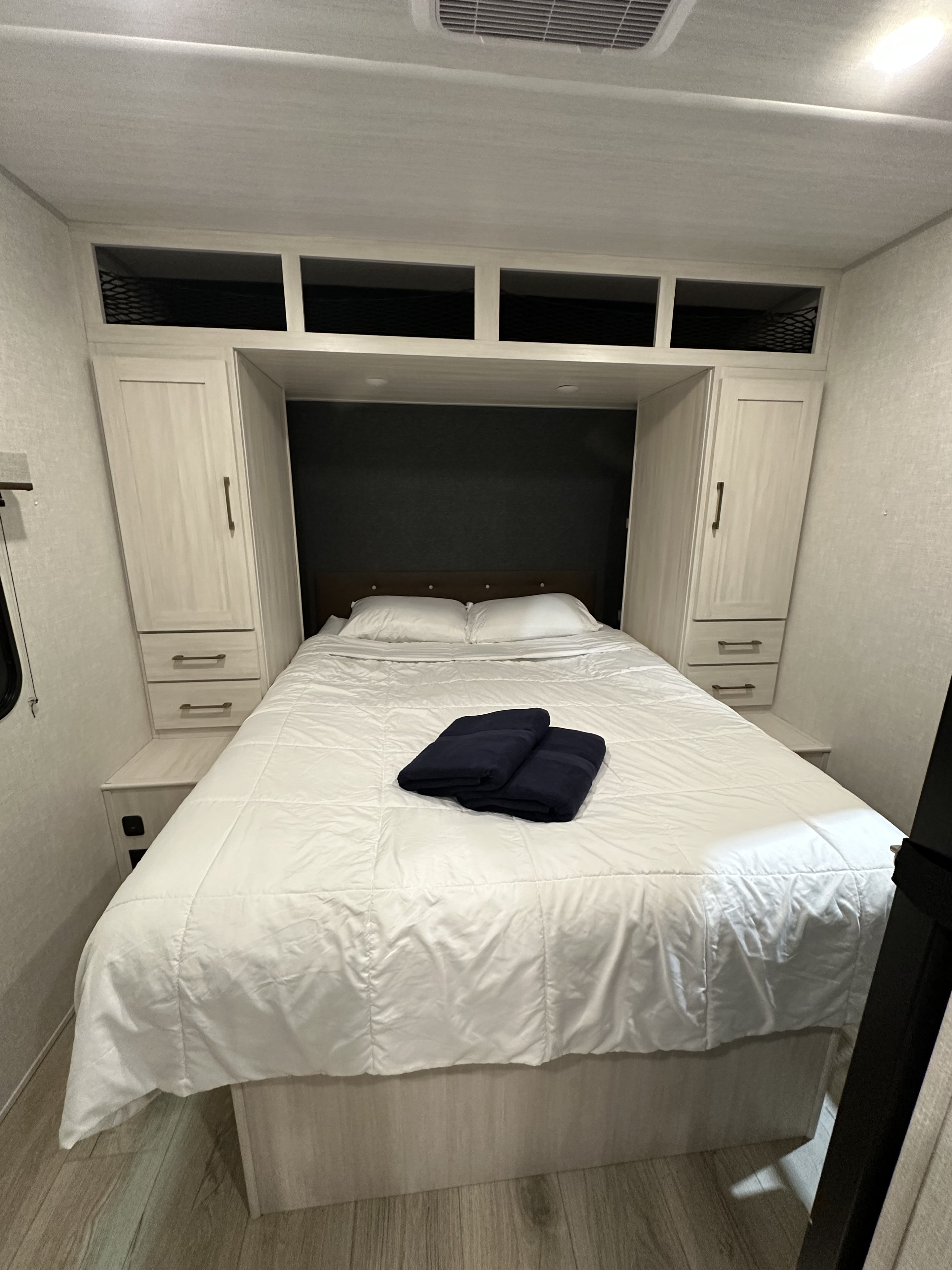 Clean RV rental bedroom featuring a plush white bed, dark headboard, light wood cabinets, and overhead storage.. Forest River Surveyor 2026