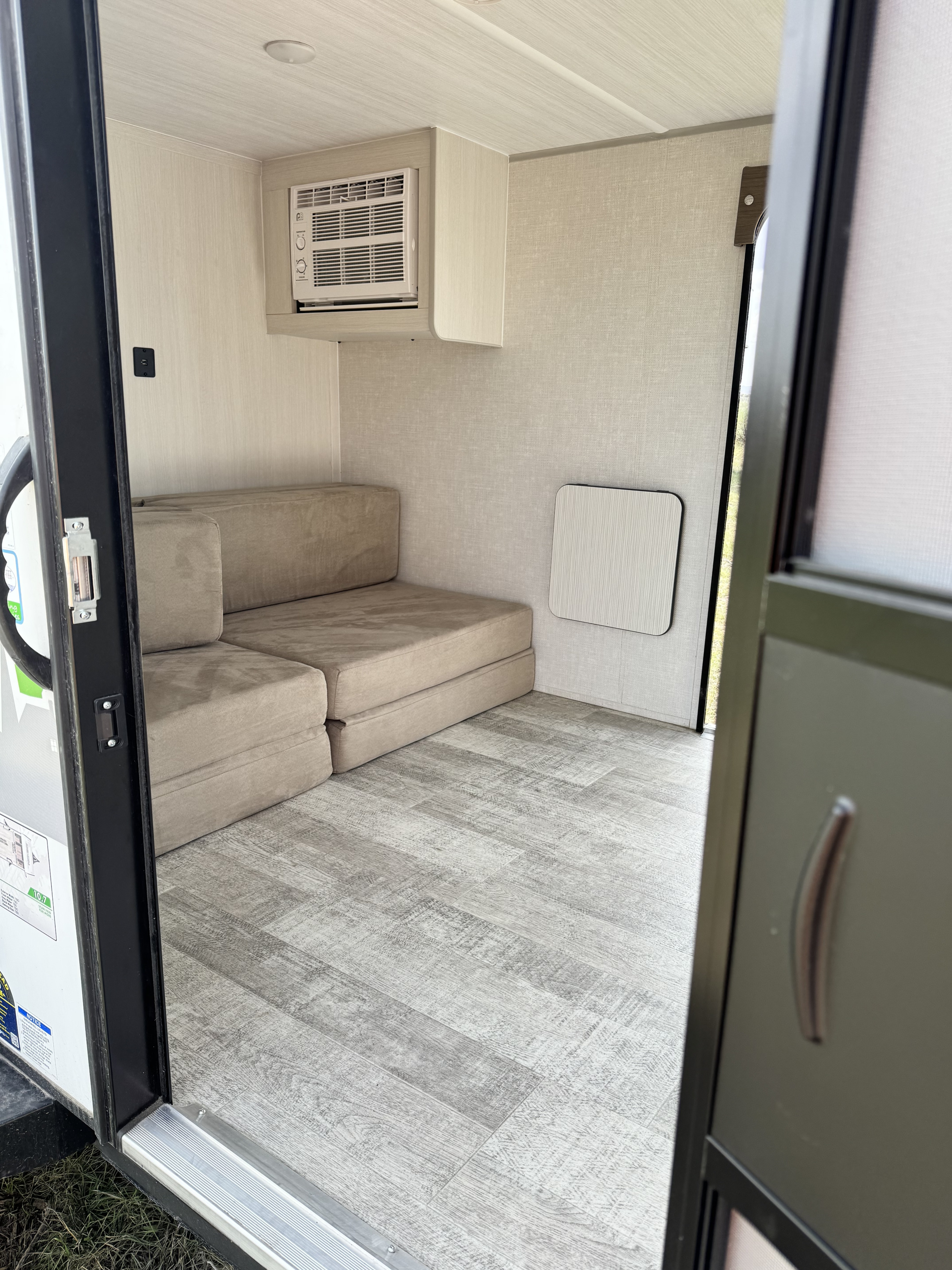 Inside an RV rental, showing cozy modular seating, a window AC unit, light wood-look flooring, and a wall table.. No Boundaries 16 2022