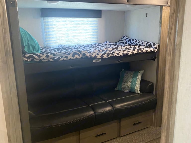 Cozy RV rental interior: a top bunk with patterned bedding, a black leather-look sofa below, and storage drawers. Ideal for travel.. Forest River Cherokee Arctic Wolf 2021