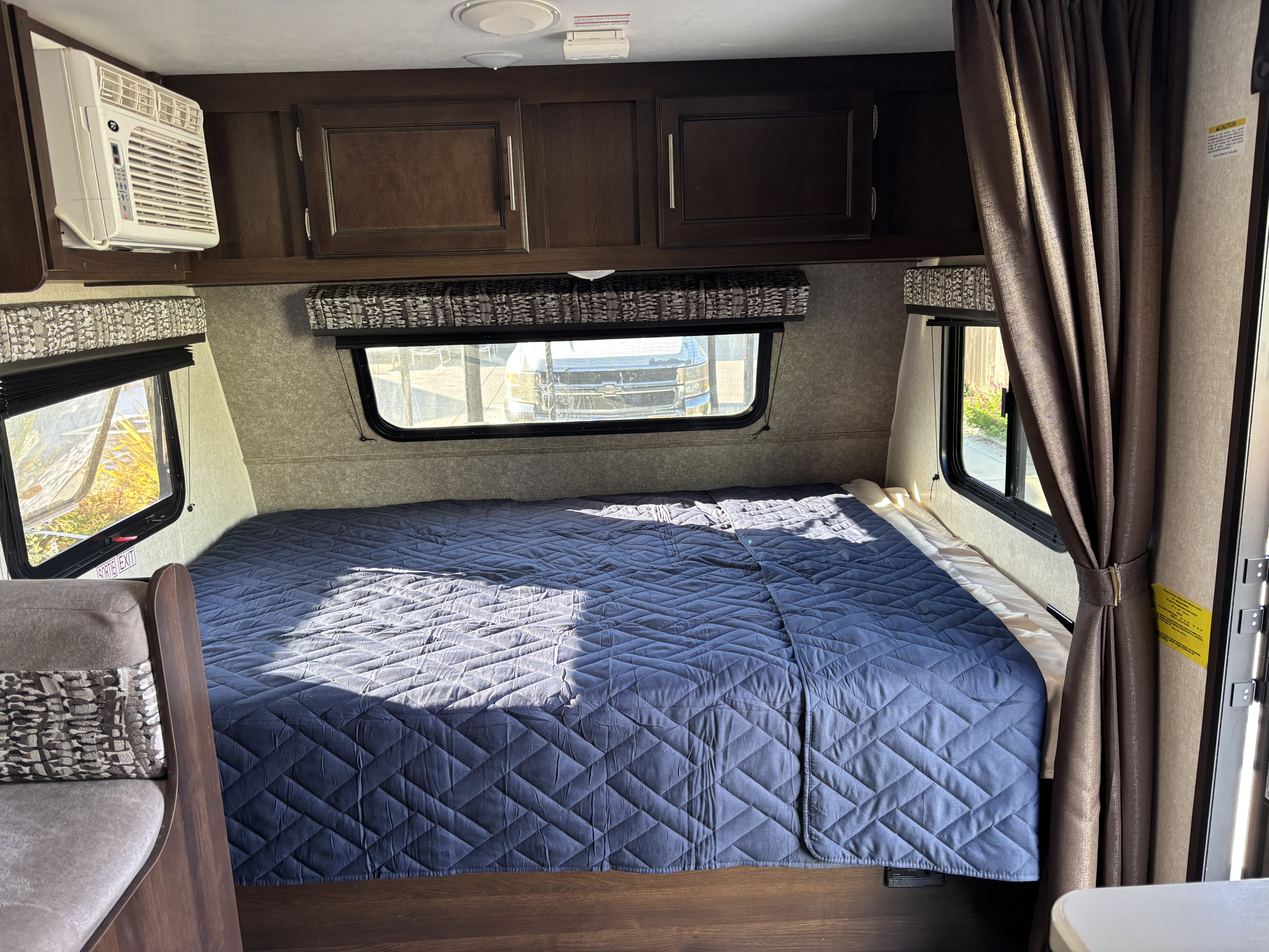 RV rental interior: cozy bedroom with blue quilted bed, overhead cabinets, windows, and an air conditioning unit.. Jayco Jay Flight SLX 2019