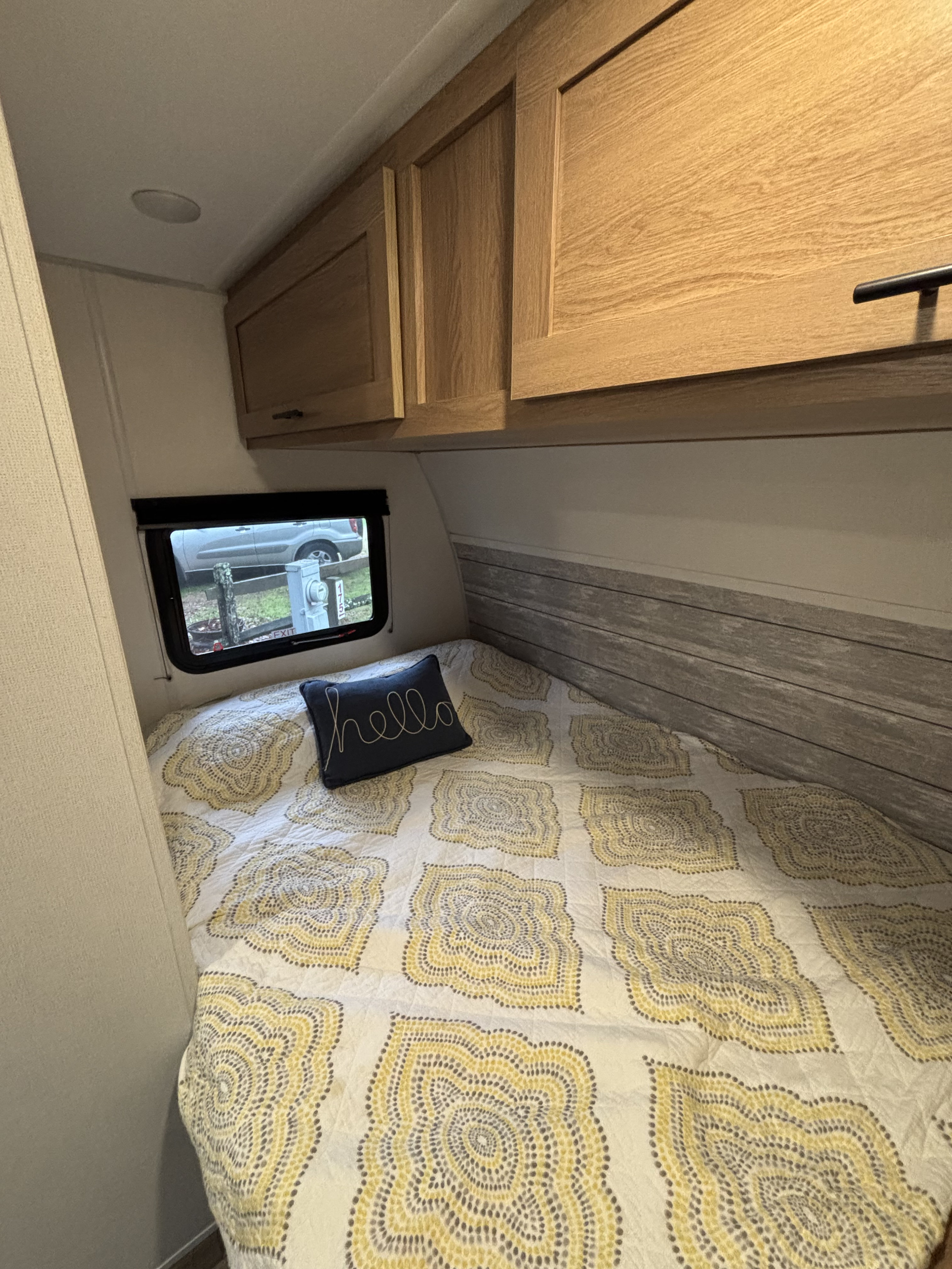 Inside a comfortable RV rental bedroom, featuring a patterned bedspread, "hello" pillow, overhead cabinets, and a campsite view.. Avenger 16RD 2024