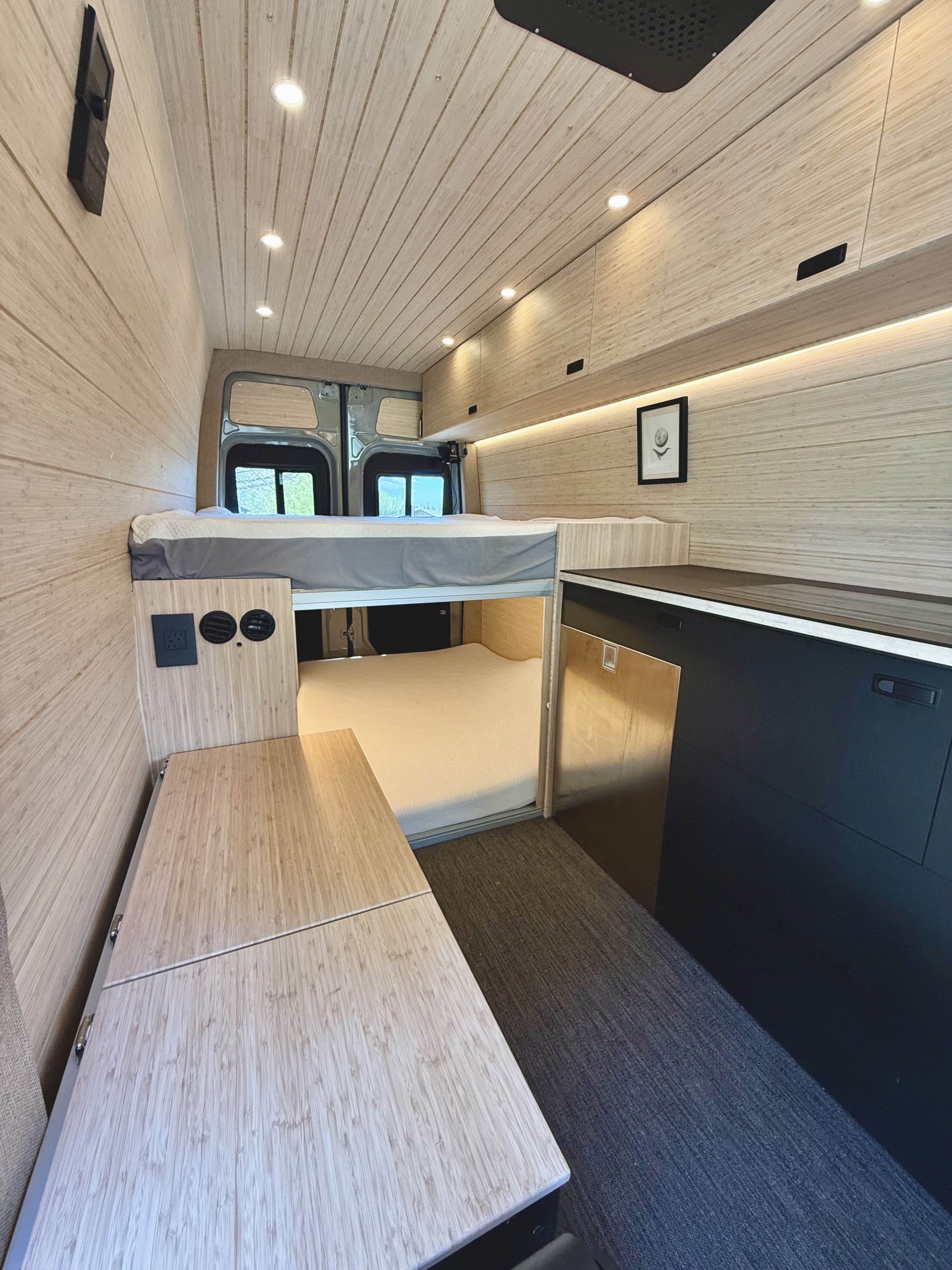RV rental featuring a sleek camper van interior with natural wood paneling, an elevated bed, and compact kitchen.. Mercedes-Benz Sprinter 2024