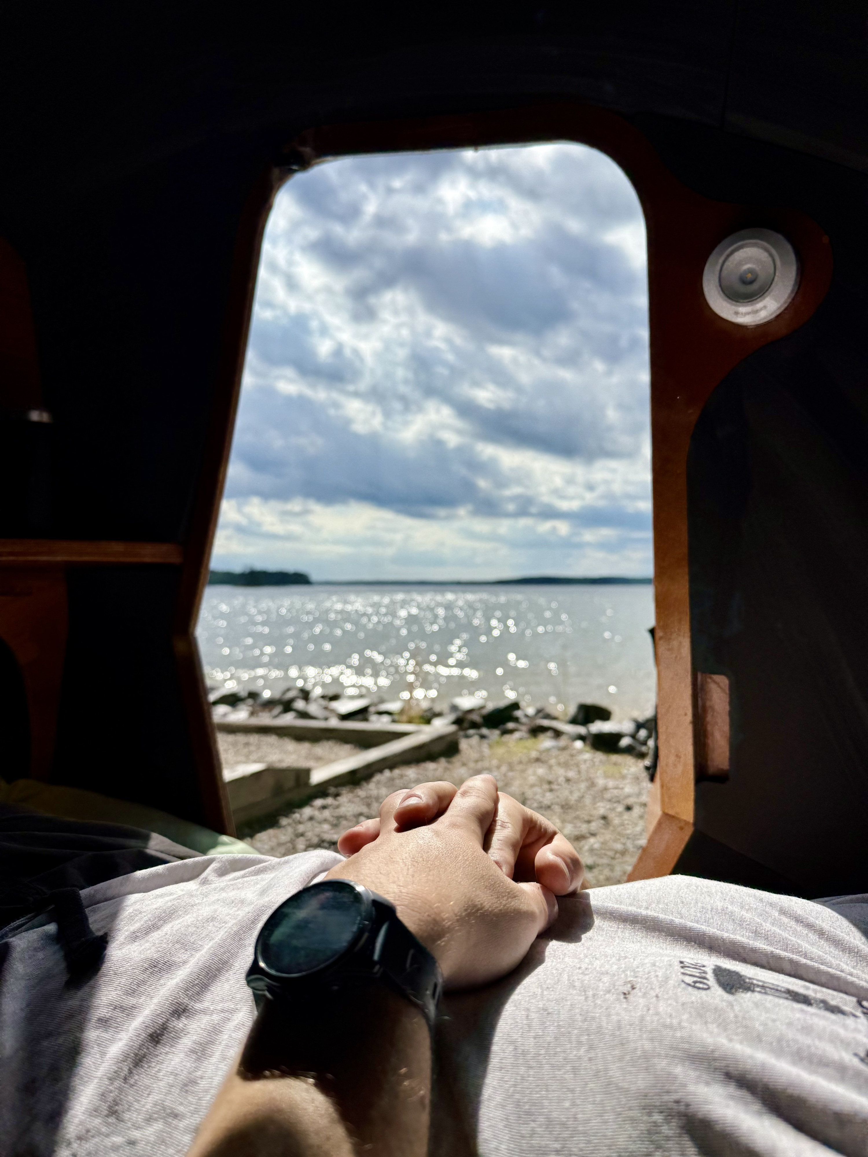 Cozy RV rental interior looking out at a sparkling lake and cloudy sky. Person's hands clasped with smartwatch.. Chesapeake Light Craft CLC Teardrop Camper 2025