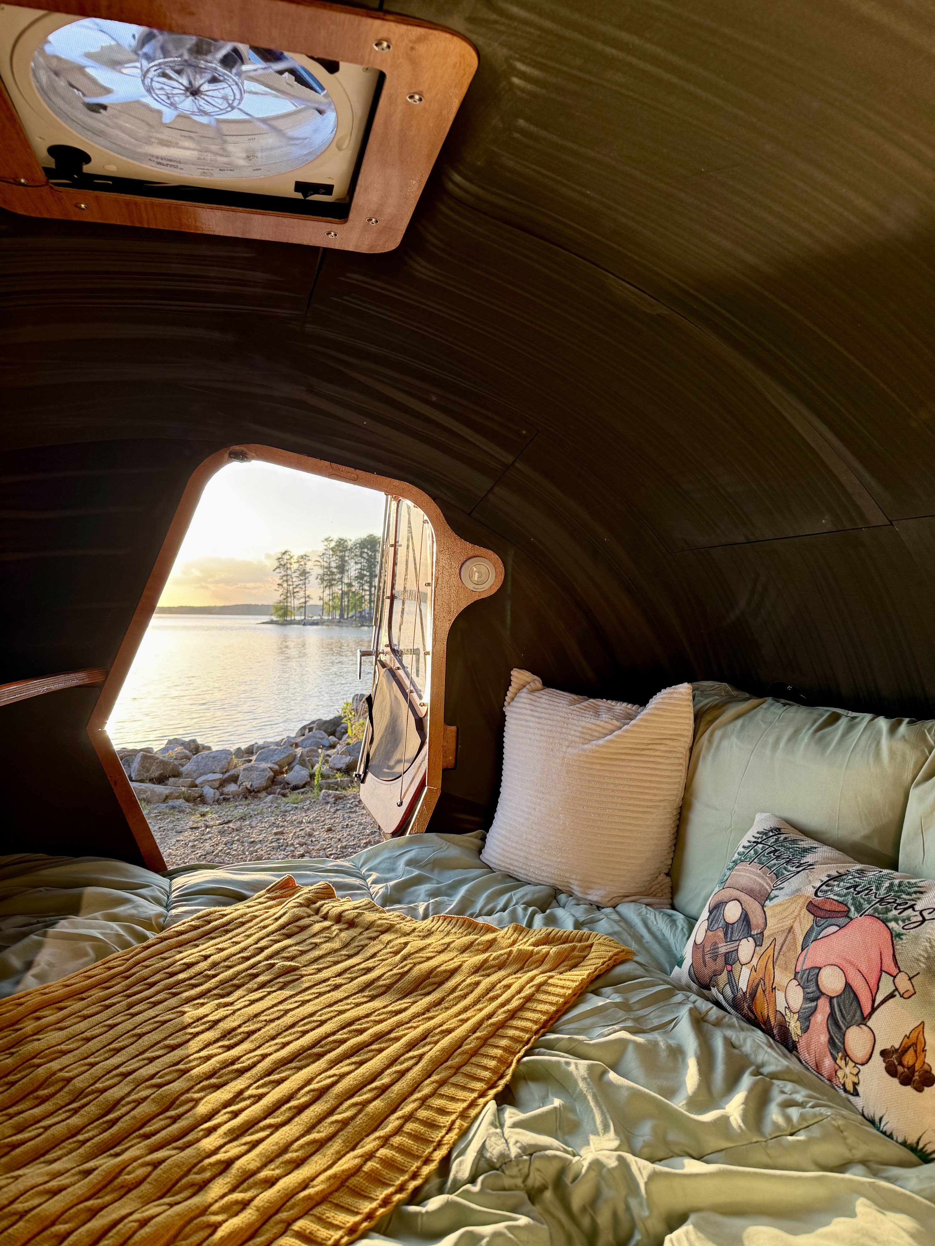 Inside a cozy RV rental, a comfy bed faces an open door with a beautiful lake & sunset view. Ideal for camping.. Chesapeake Light Craft CLC Teardrop Camper 2025