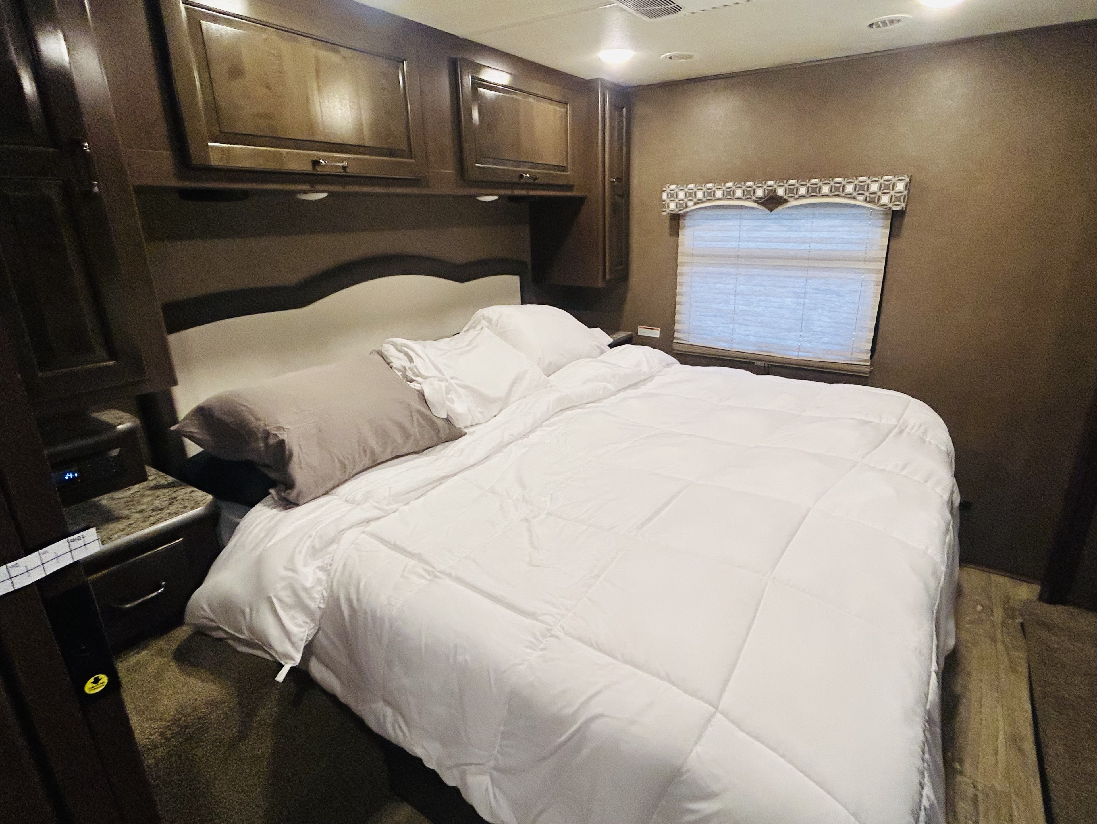 Cozy RV rental bedroom with a neatly made white bed, overhead cabinets, and a window. Perfect for travelers.. Thor Hurricane 2018