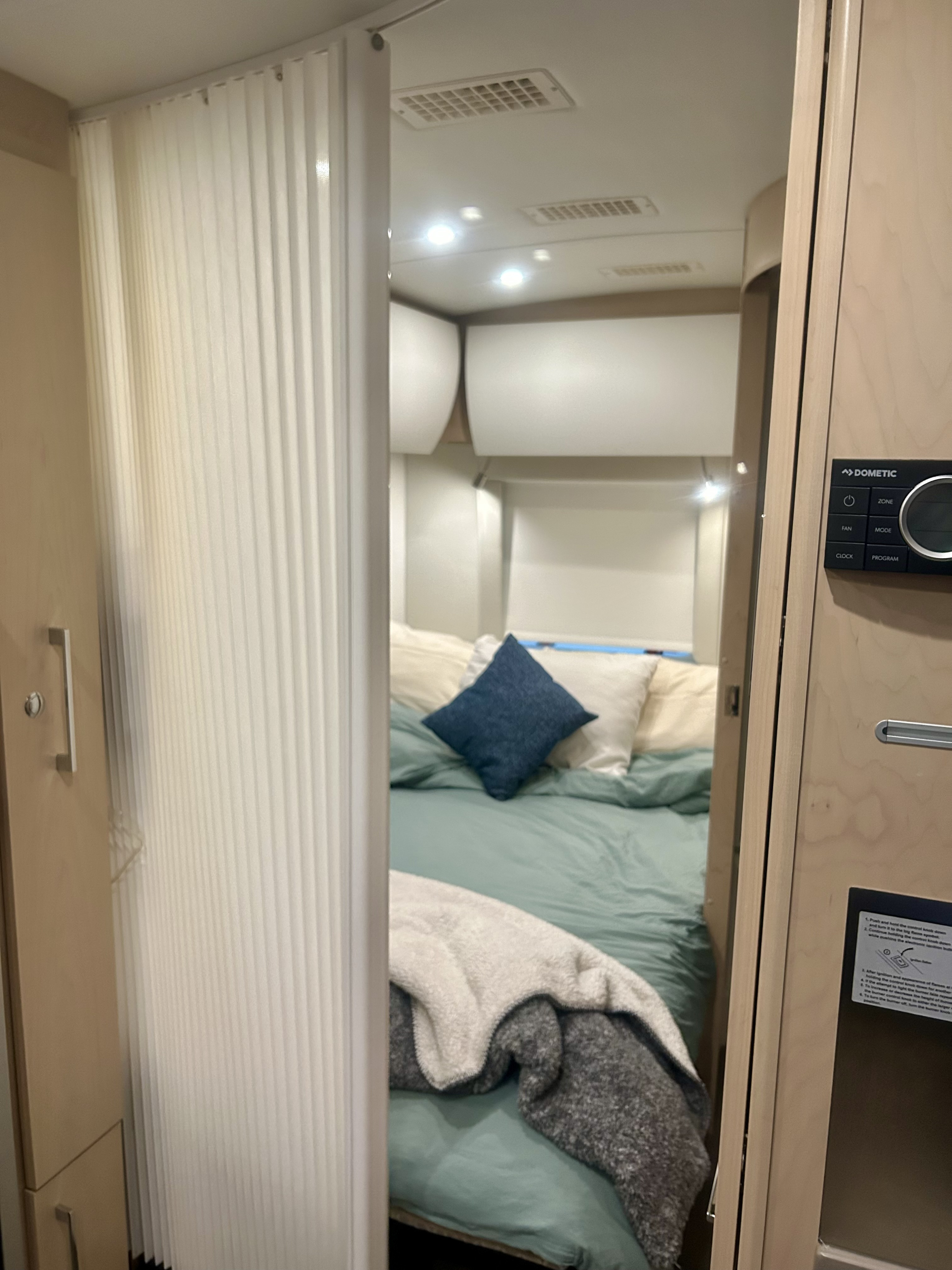 Compact RV rental interior: a peek into the bedroom with a comfortable bed, pleated room divider, and Dometic control panel.. Leisure Travel Vans Serenity 2022