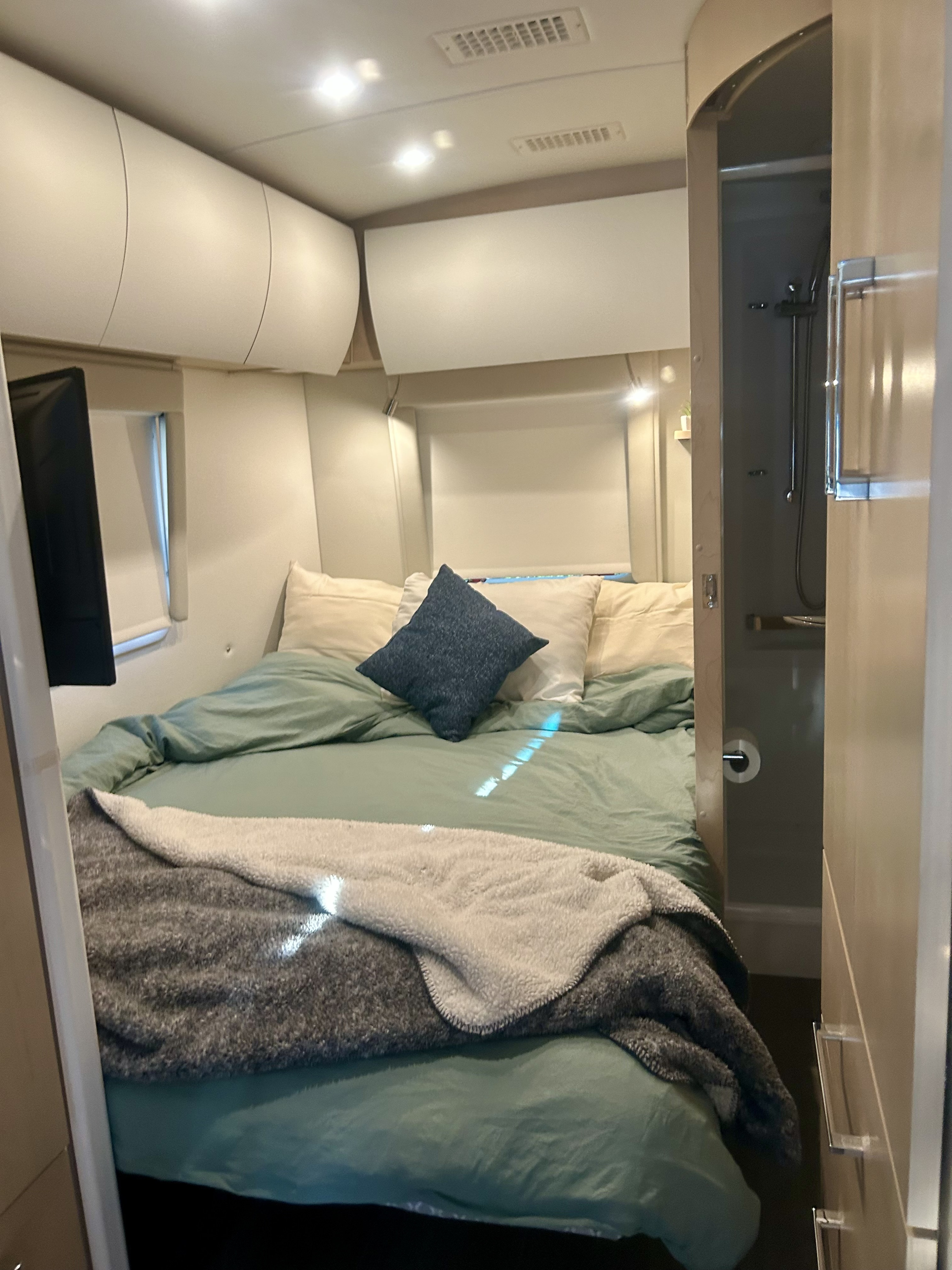 Inside a comfortable RV rental: a compact bedroom with a green-sheeted bed, overhead cabinets, and an adjacent shower.. Leisure Travel Vans Serenity 2022
