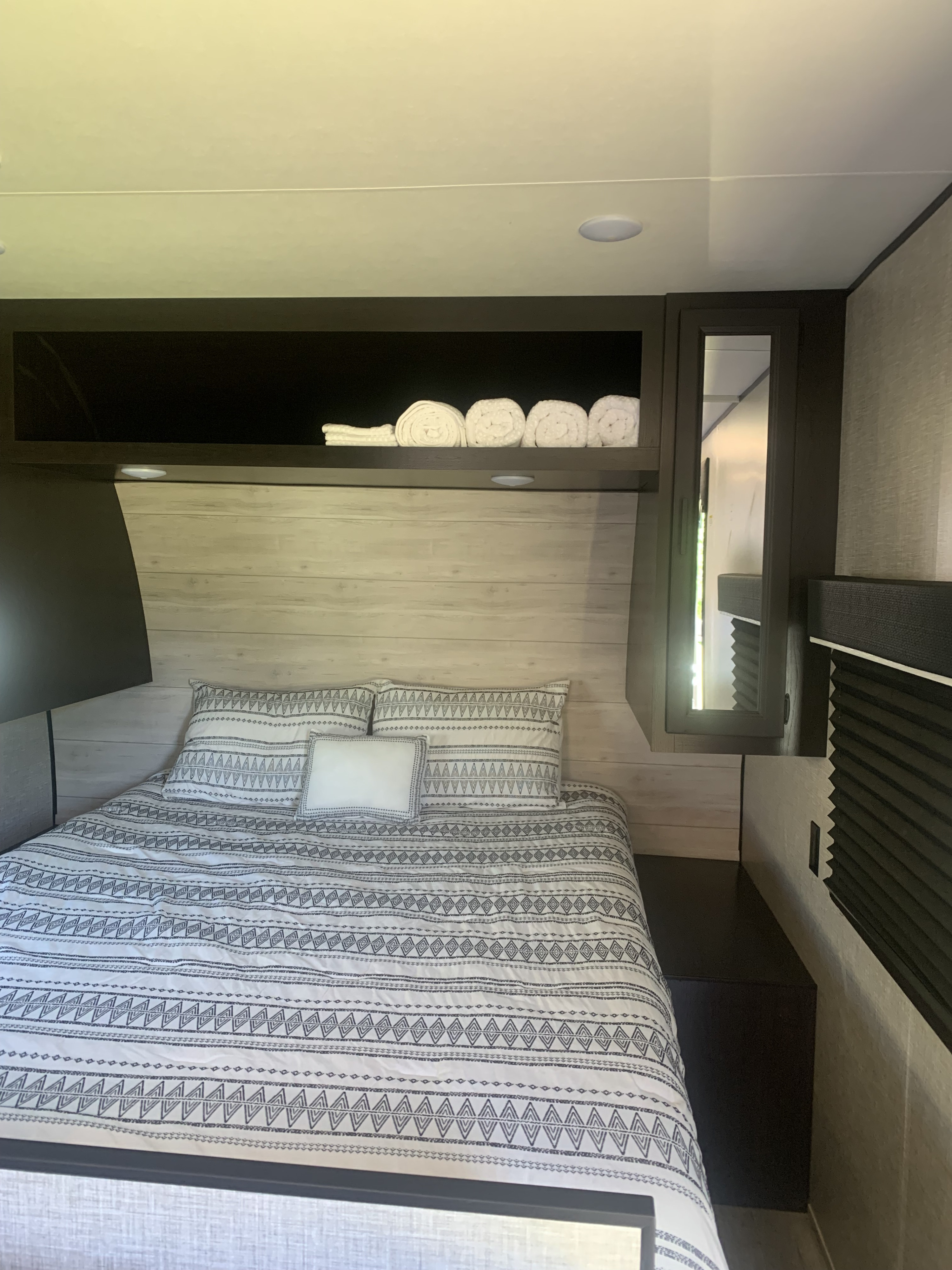 Cozy **rv rental** bedroom: patterned bed, light wood paneling, overhead storage with towels, and a mirrored cabinet.. Jayco Jay Flight 2023