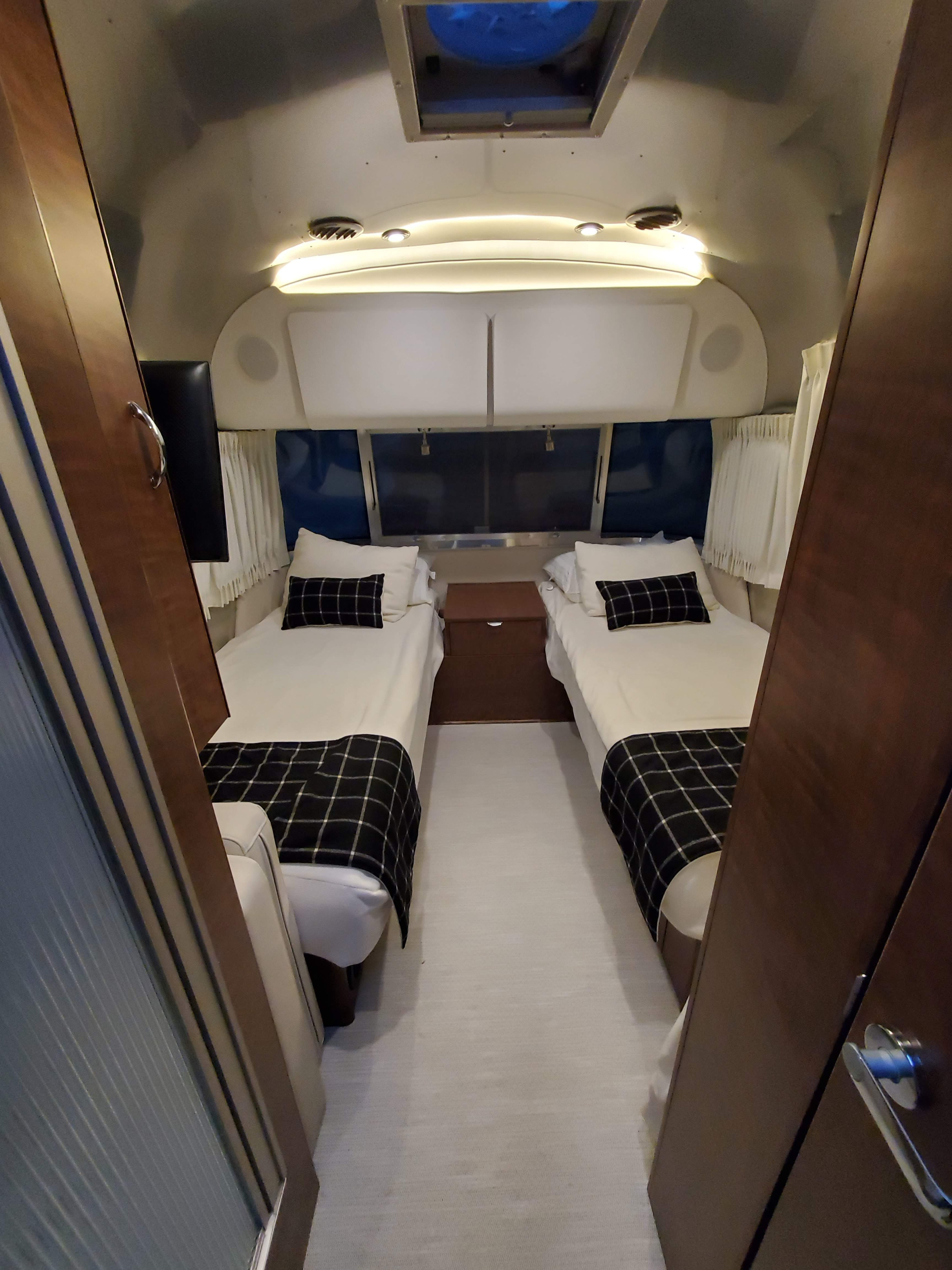 Explore this RV rental's cozy bedroom: twin beds with plaid blankets, a nightstand, & built-in TV for your travels.. Airstream Globetrotter 2019