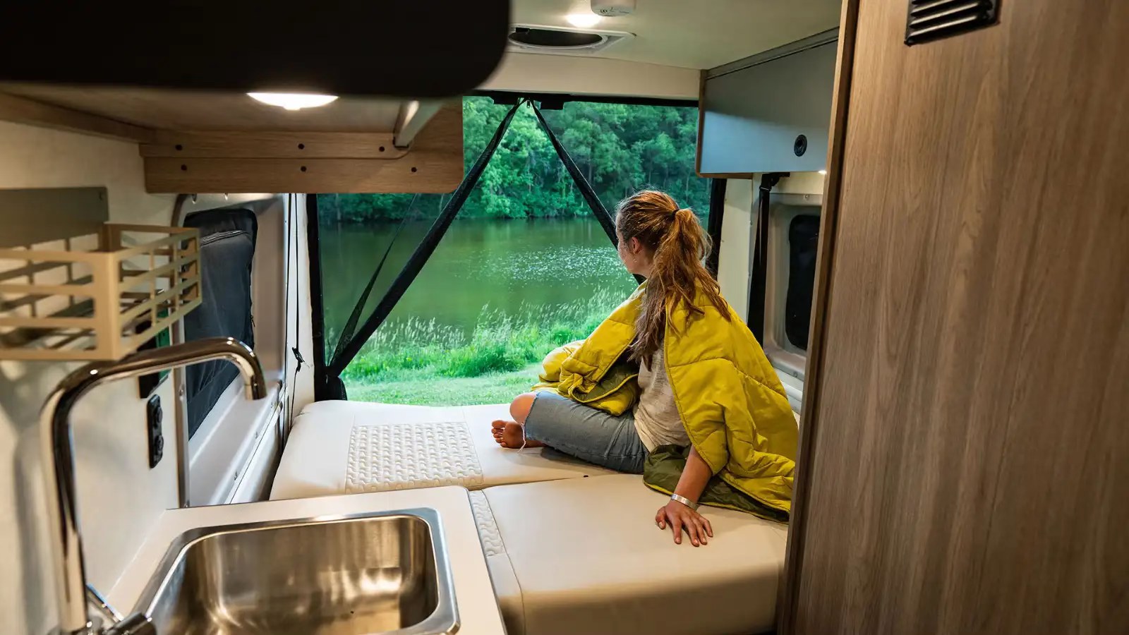 RV rental: Woman enjoys a serene lake view from inside her cozy camper van, equipped with a kitchen sink.. Winnebago Solis 2025