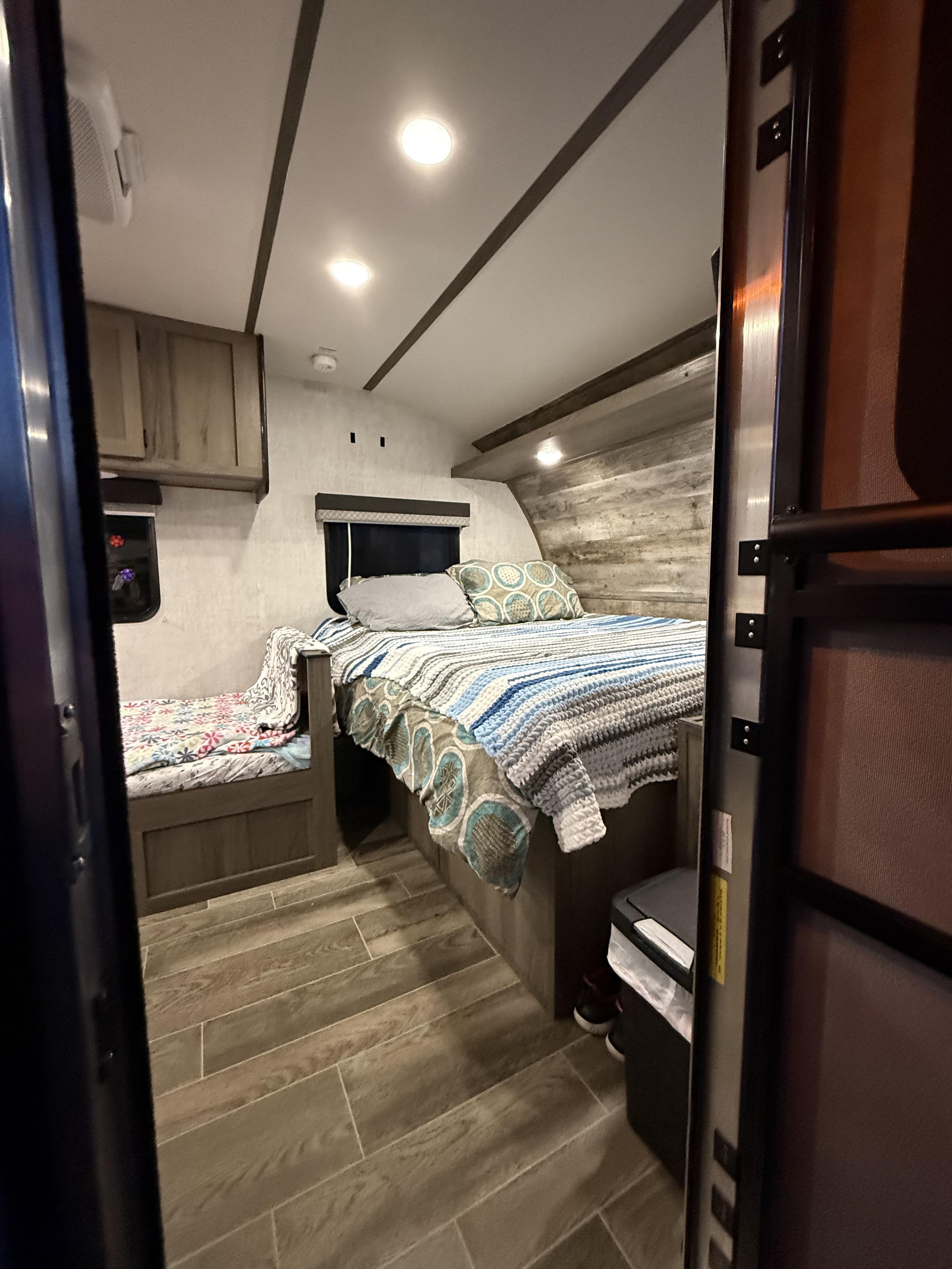 Inside a bright RV rental: cozy main bed, bunk bed with colorful duvet, wood-look floors, and ceiling lights.. Gulf Stream Enlighten 2022