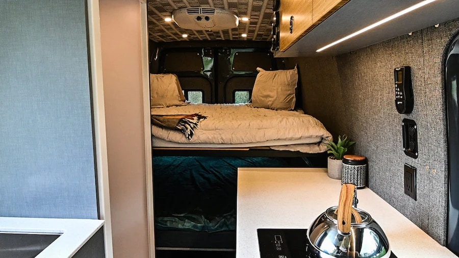 Cozy RV rental interior with a raised bed, lower bunk space, and a compact kitchen counter with kettle.. Mercedes-Benz Sprinter 2021
