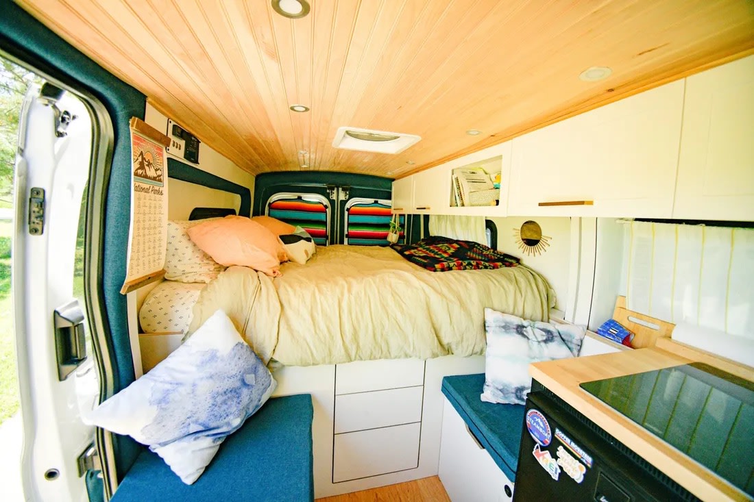 Inside a cozy RV rental: a comfortable bed at the back, wood-paneled ceiling, storage, and a compact kitchen area.. Ford Transit 2019