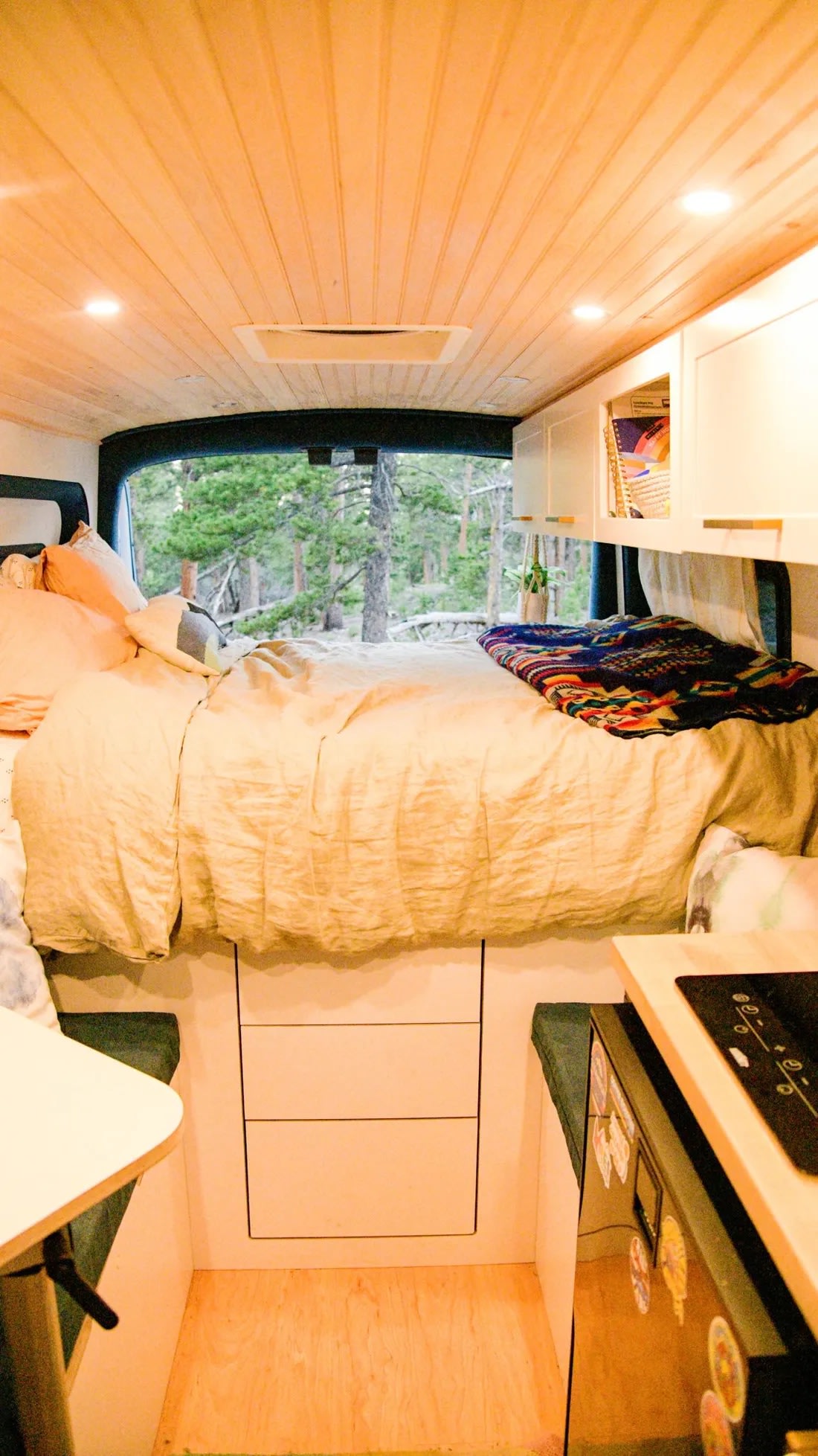 RV rental camper van interior. Features cozy bed looking out to forest, kitchenette, and wood-paneled ceiling.. Ford Transit 2019