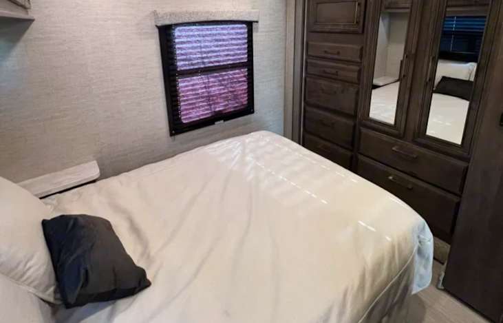 RV rental bedroom with a white bed, window with purple blinds, and dark wood mirrored cabinet.. Entegra Coach Odyssey 31F 2022