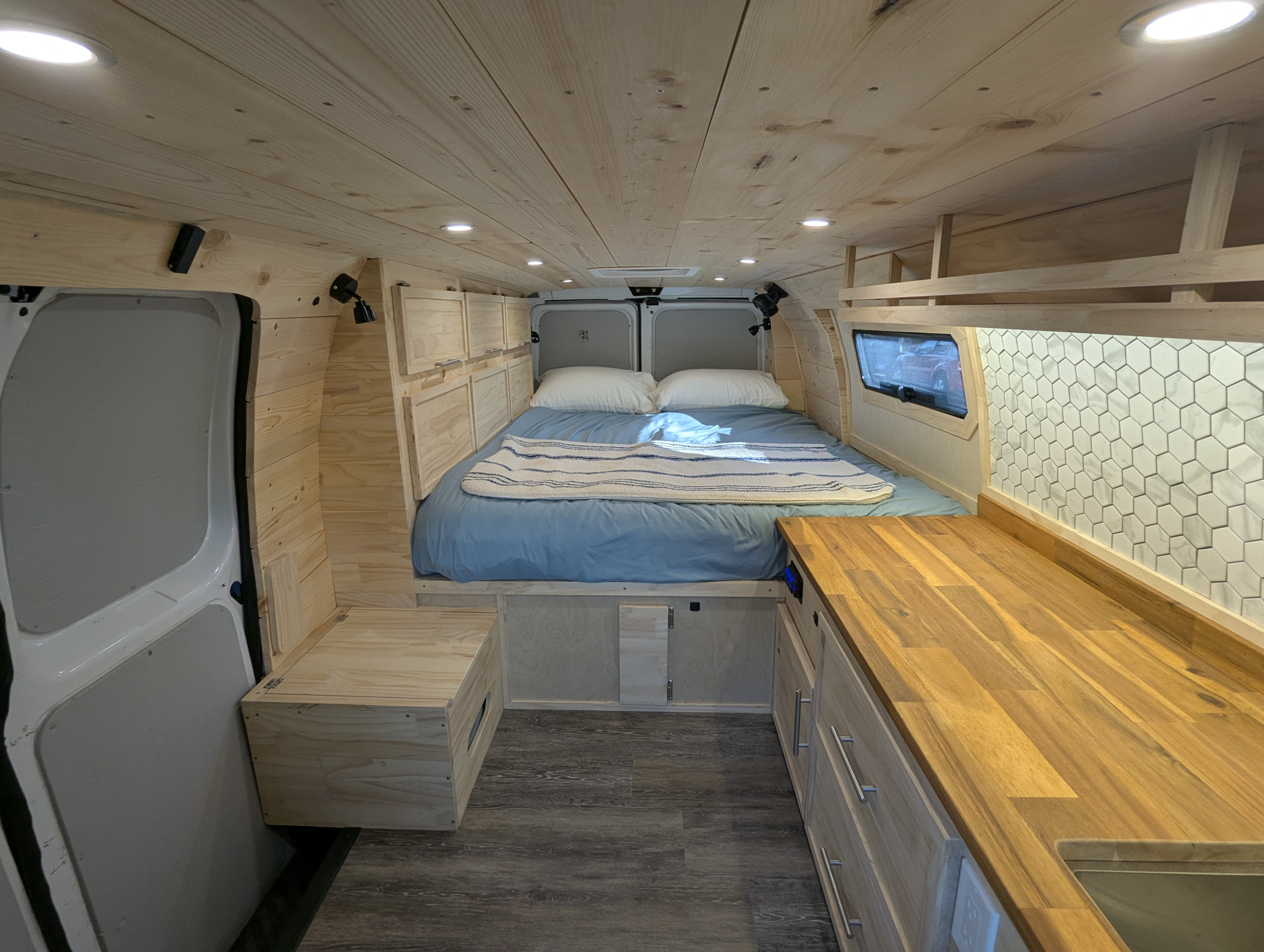 Inside a custom wood-lined camper van for RV rental, featuring a cozy bed, countertop, and built-in storage.. Gmc Savanna 2016