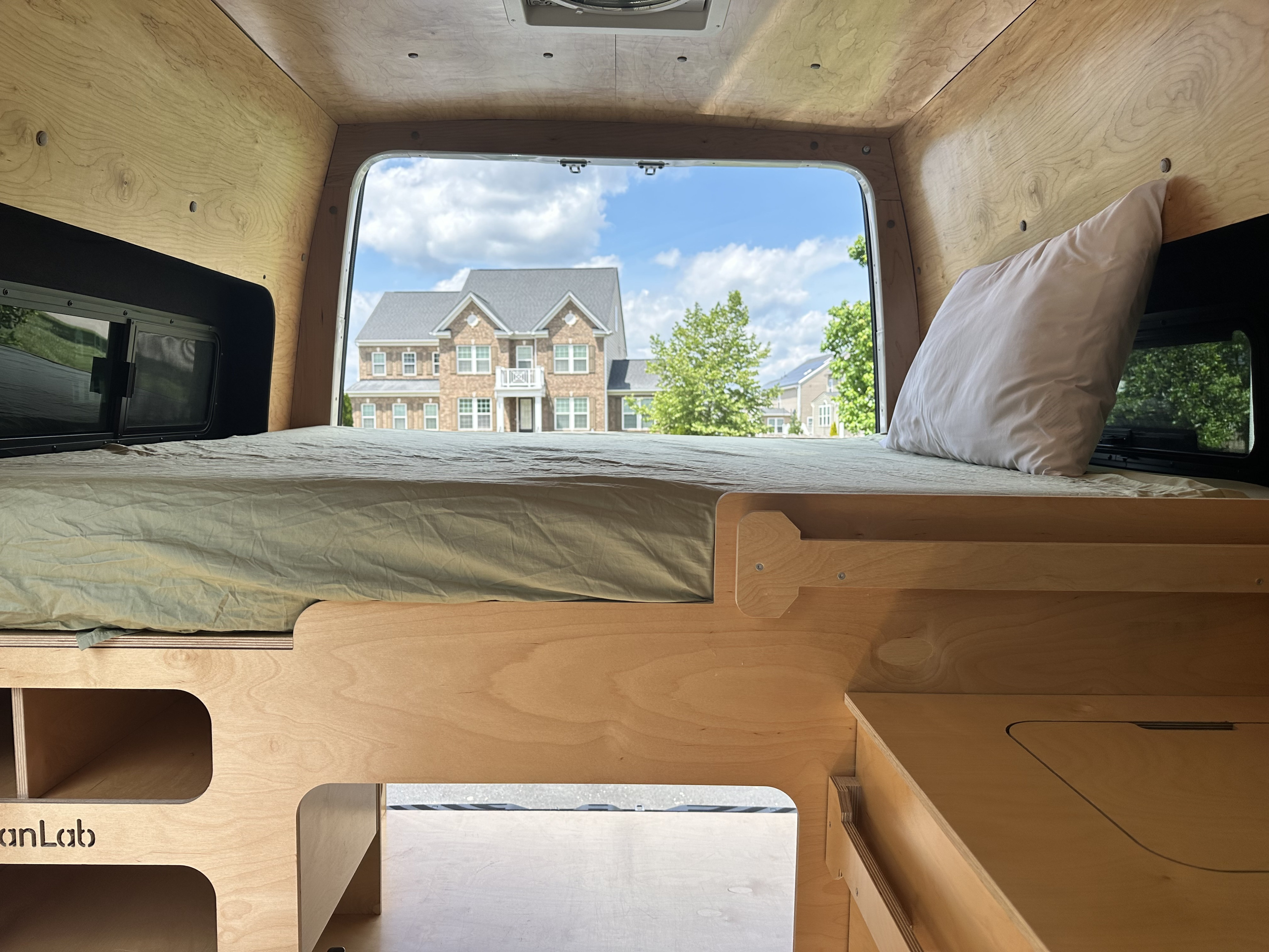 RV rental interior: a comfy bed within a wood-paneled camper van, looking out at a house through the rear window.. Mercedes-Benz Sprinter 2021