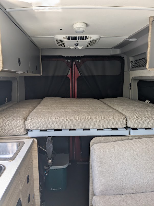 Inside an RV rental: a cozy bed over a kitchen area, living space, and rear doors. Includes overhead storage, AC, and a cooler.. Winnebago Solis 2023