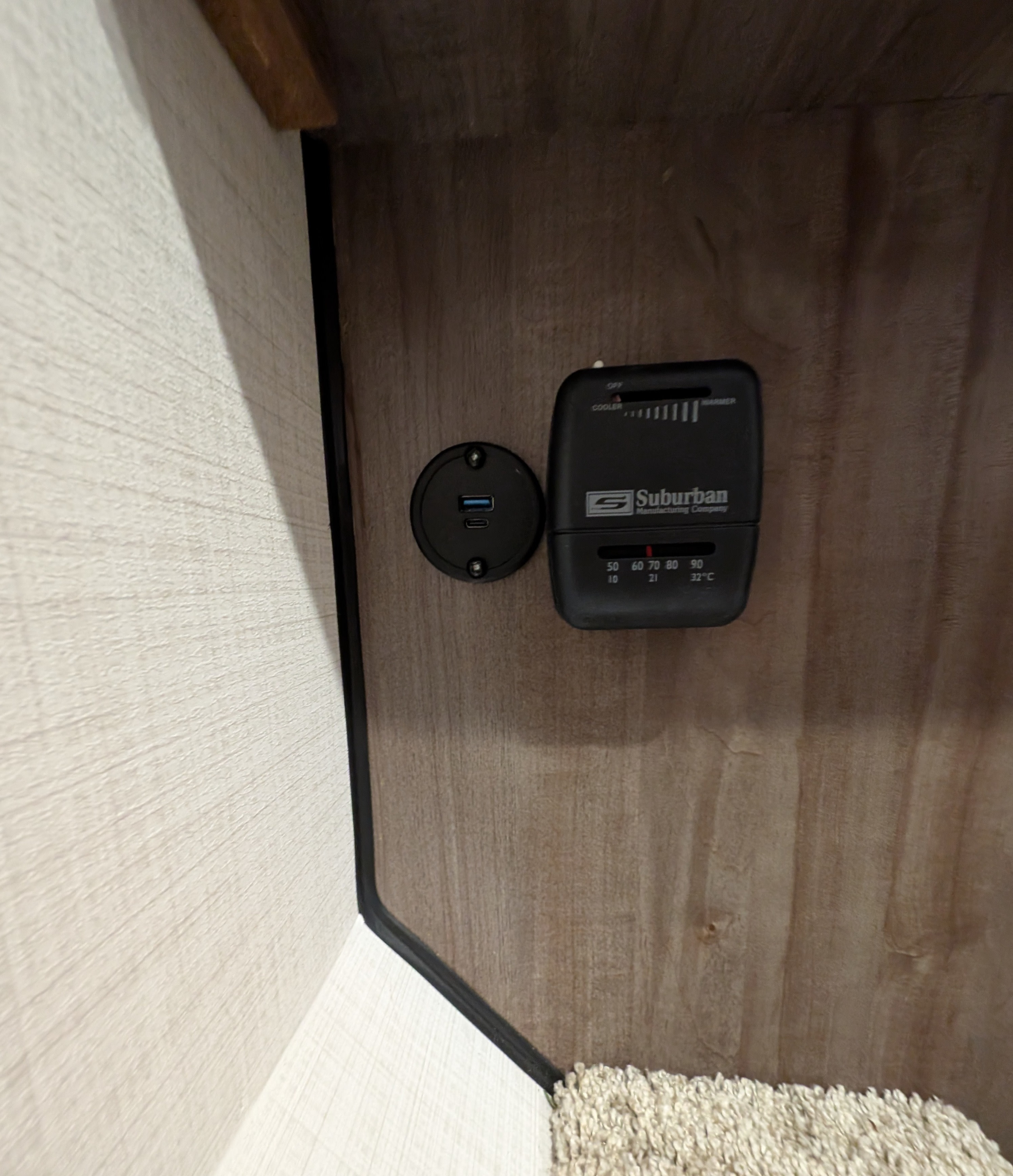 RV rental controls: A Suburban thermostat and USB charging station are mounted on a wood panel wall, with carpet below.. Forest River Rockwood LTD 2024