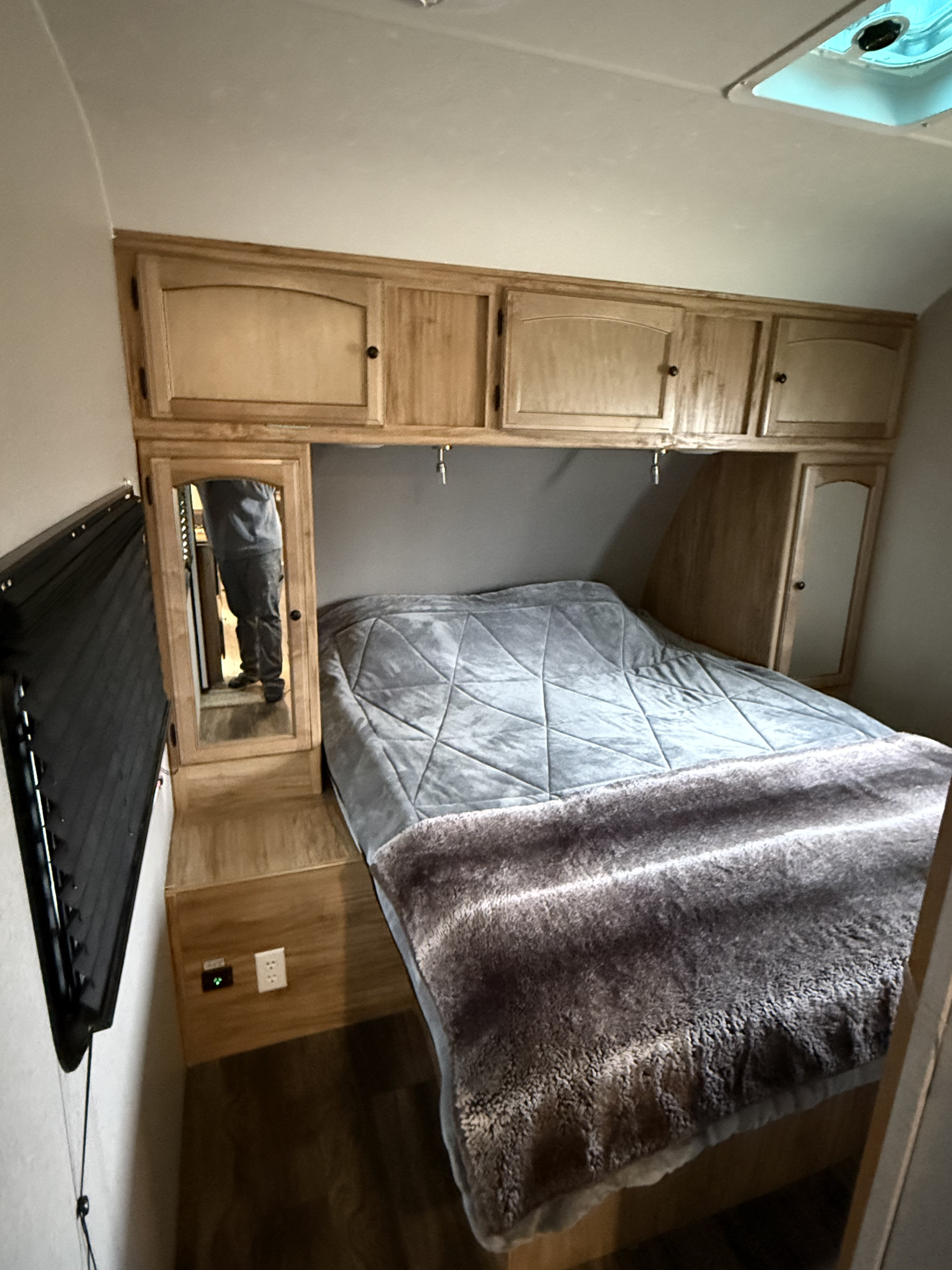 Inside a cozy RV rental bedroom: bed with grey blankets, wooden overhead cabinets, window blinds, & mirror reflecting the photographer.. Coachmen Freedom express ultra light 2019