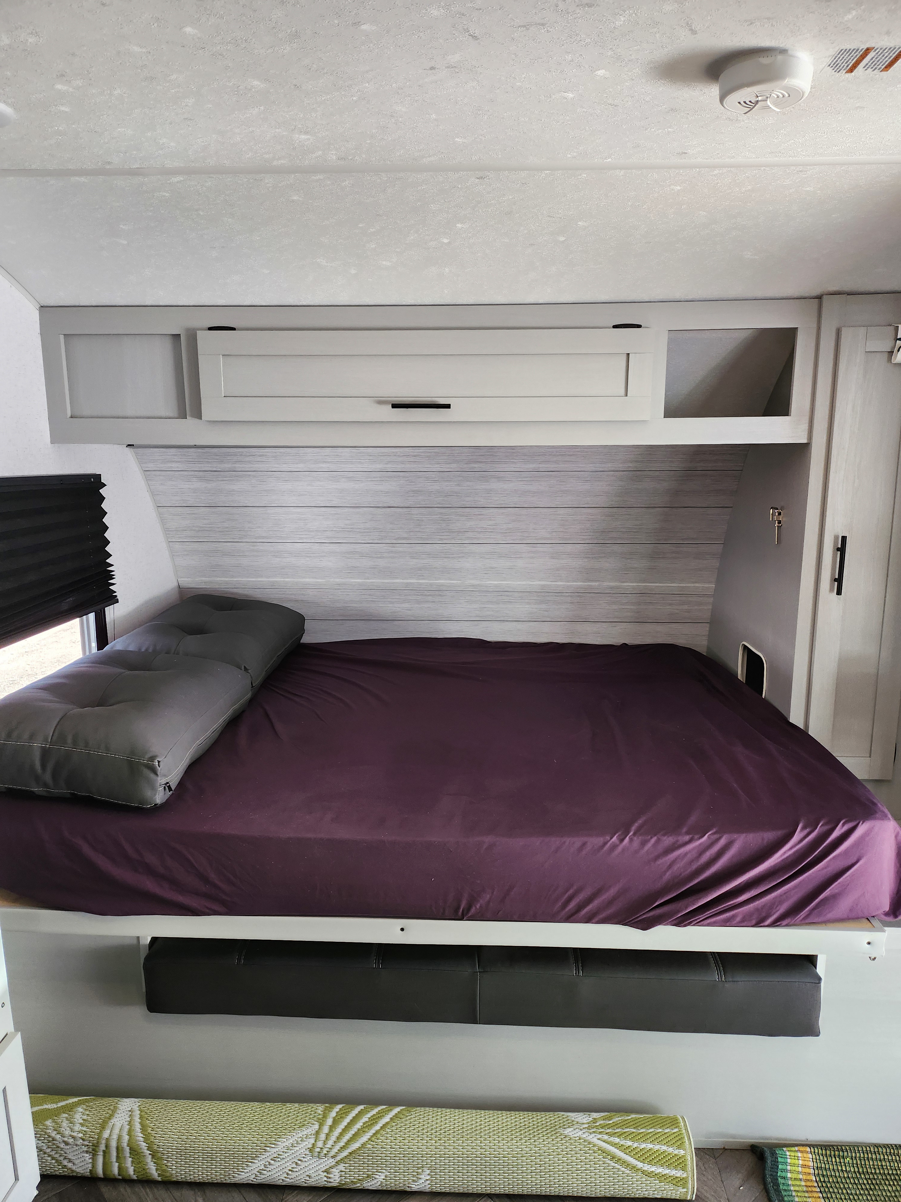 RV rental bedroom: purple bed, gray cushion, light wood walls, overhead cabinets, under-bed storage, and rolled mats below.. Forest River Wildwood FSX 2022