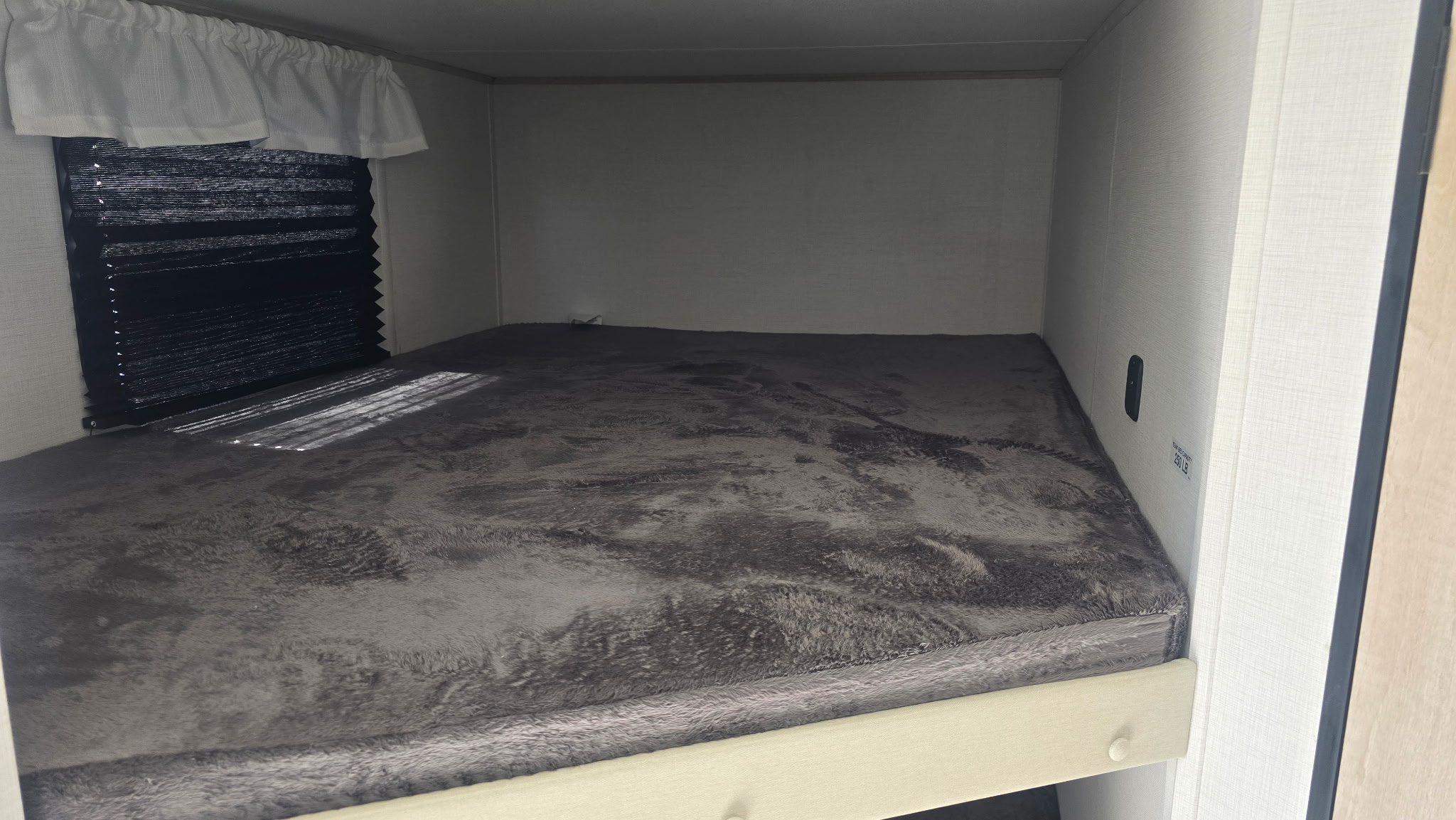 Inside your RV rental: a comfy bunk bed with a soft grey blanket, window with black pleated blind, and sun streaks.. Primetime Avenger 2025