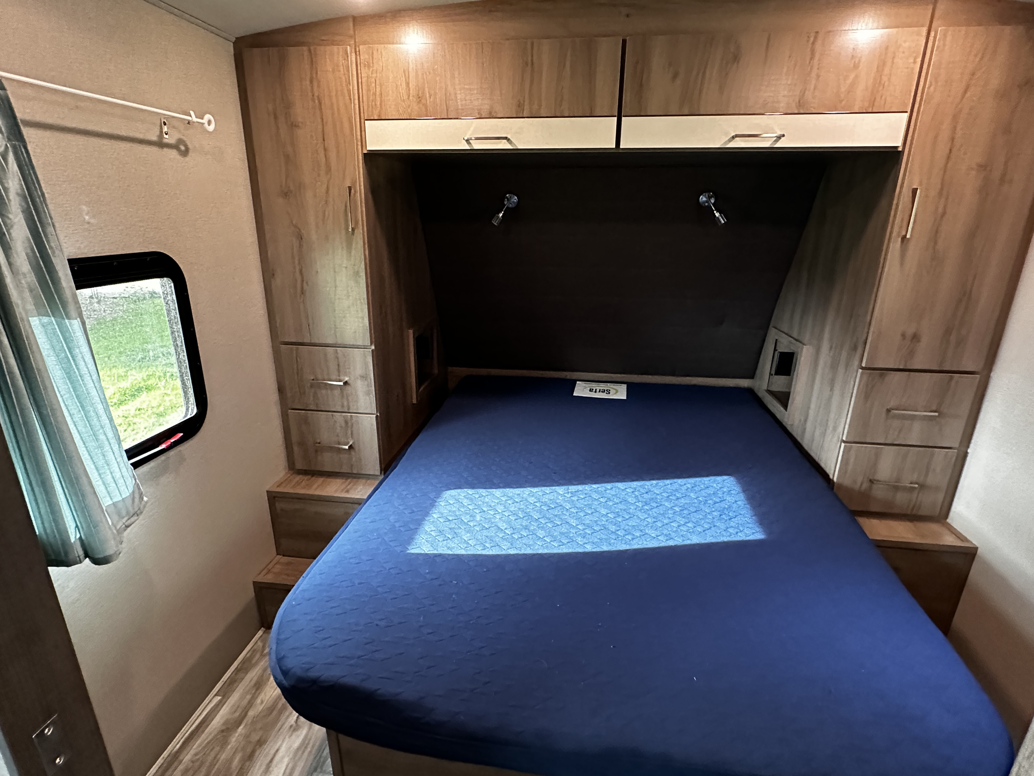 This **RV rental** bedroom features a blue bed, wooden overhead cabinets, and side storage with a window.. Grand Design Imagine 2022